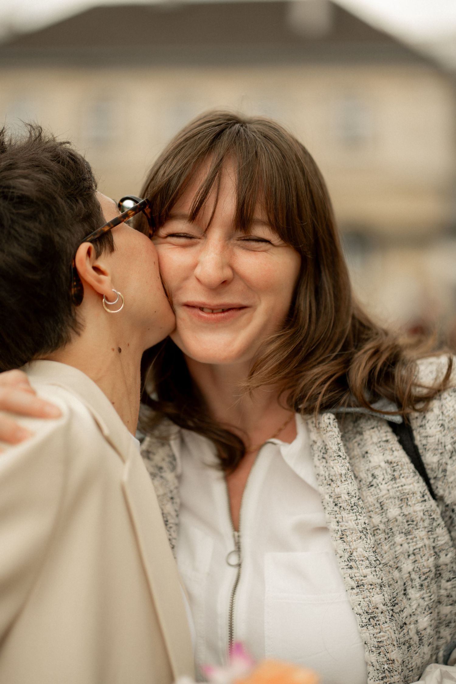 Two women sharing a tender cheek kiss outdoors, one smiling warmly in a tweed jacket at a celebration.