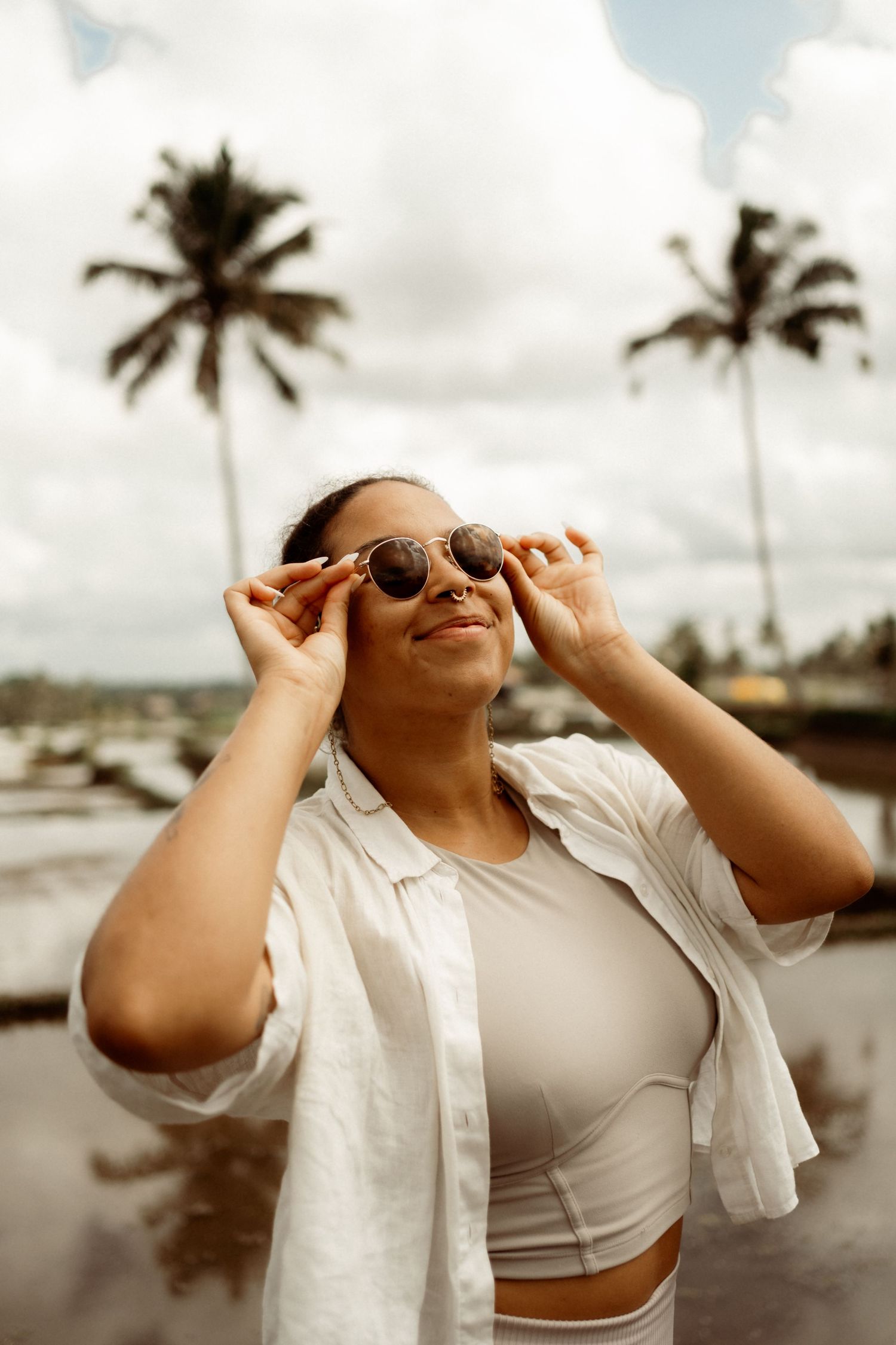 A woman adjusts her round sunglasses while standing near palm trees under a cloudy sky.