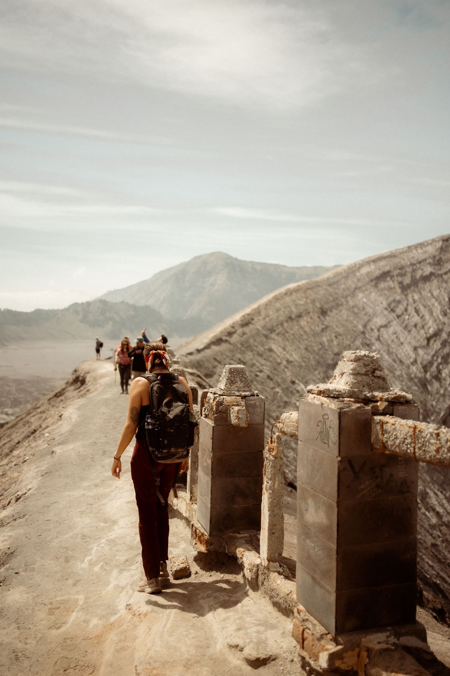 Hikers walk along a stone path on the rim of Mount Bromo volcano with misty mountains in the background.