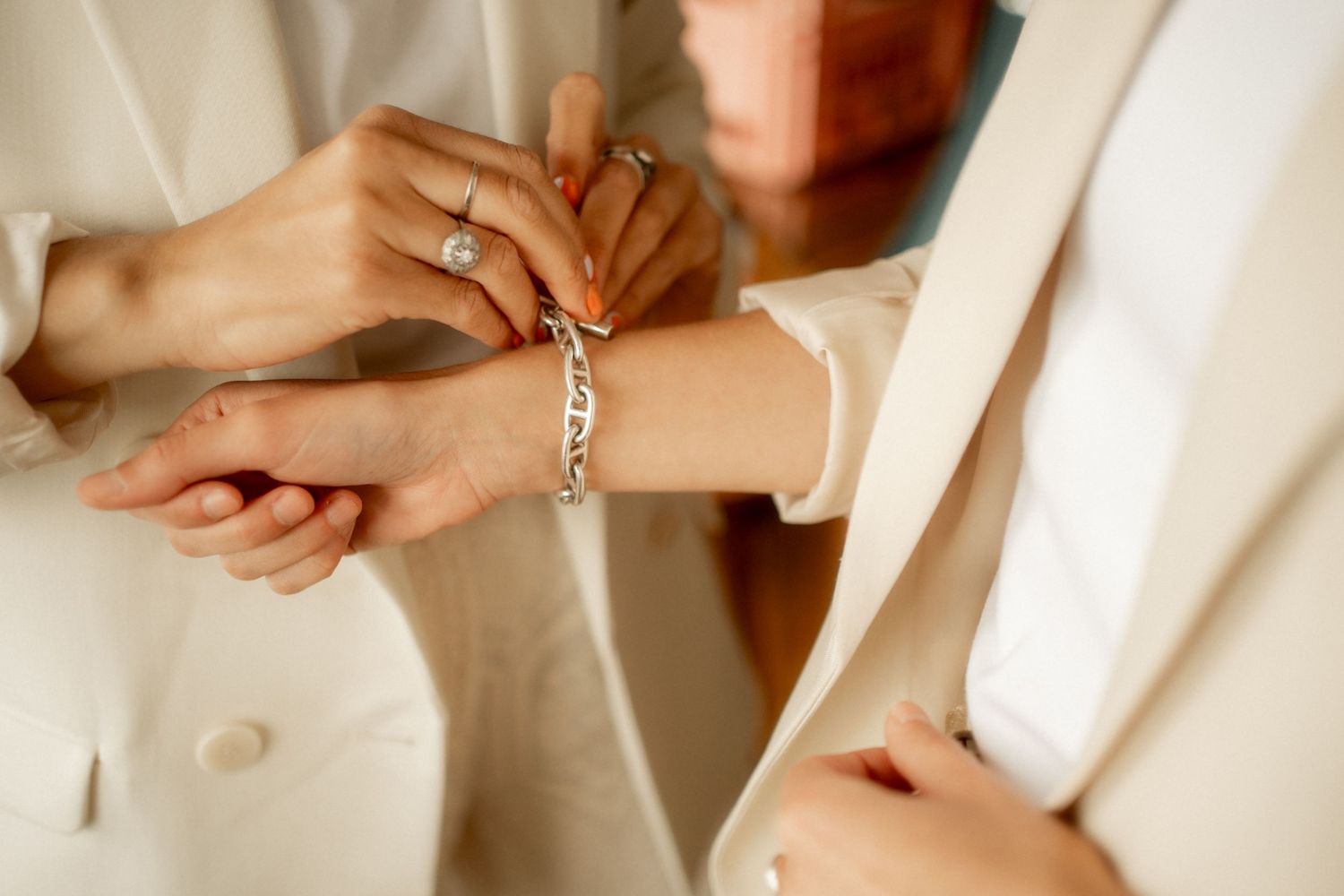 Two women in white blazers clasp a silver chain bracelet on a wrist adorned with a diamond ring and orange nail polish.