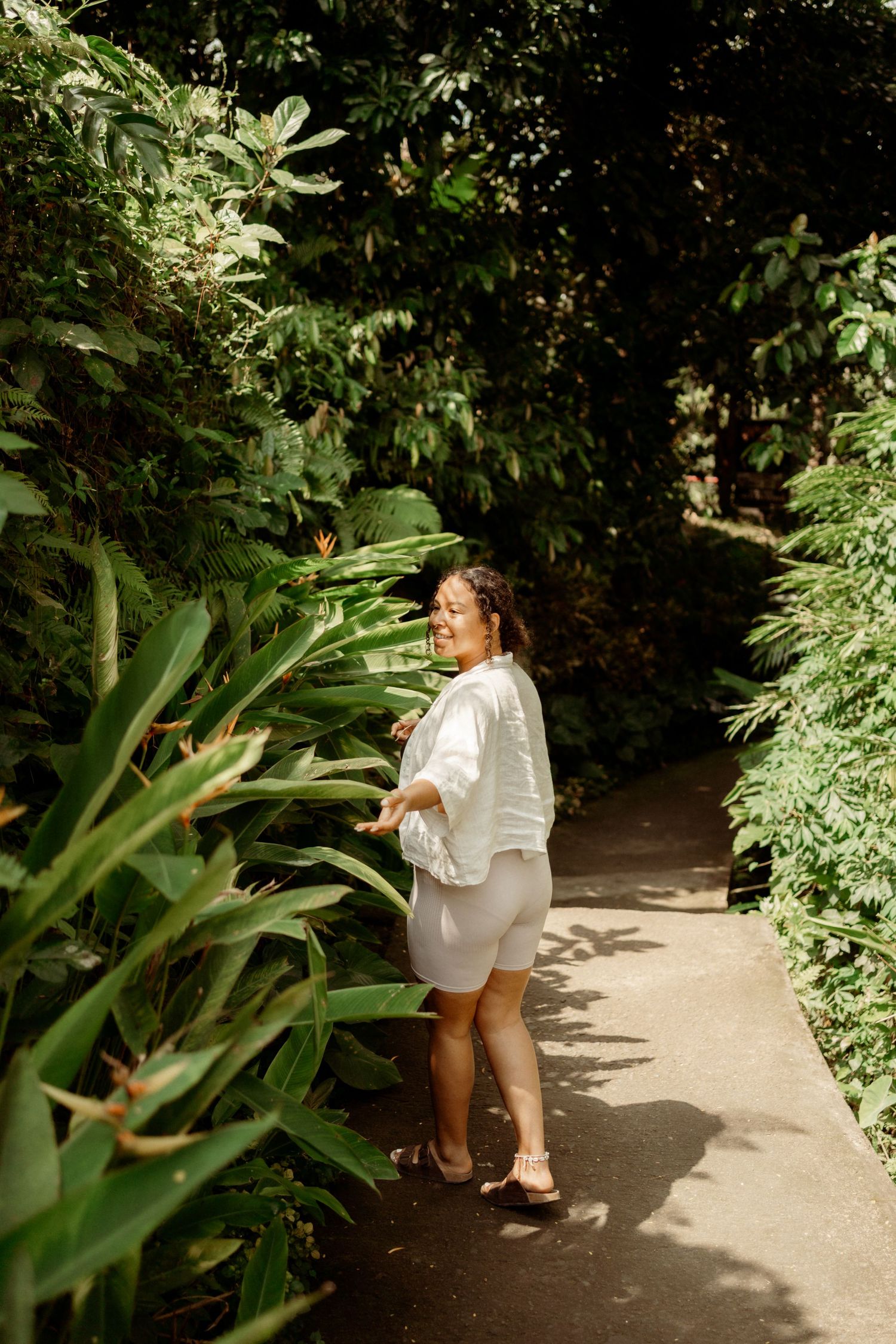 Woman in white shirt and shorts standing on a lush tropical garden path surrounded by green plants.