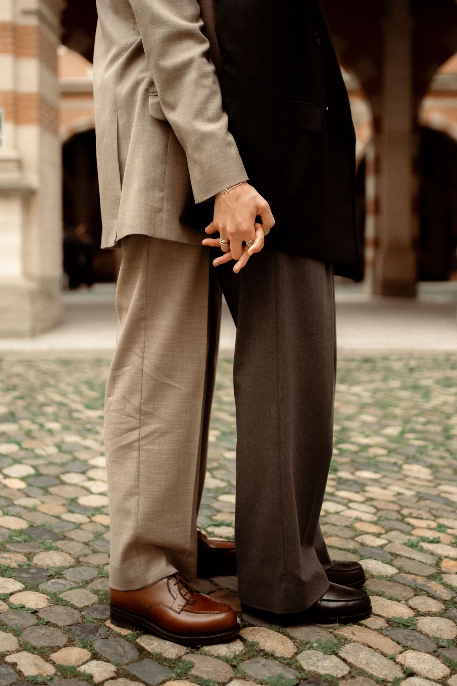 Two men in elegant suits holding hands on cobblestone courtyard, close-up of legs and shoes.