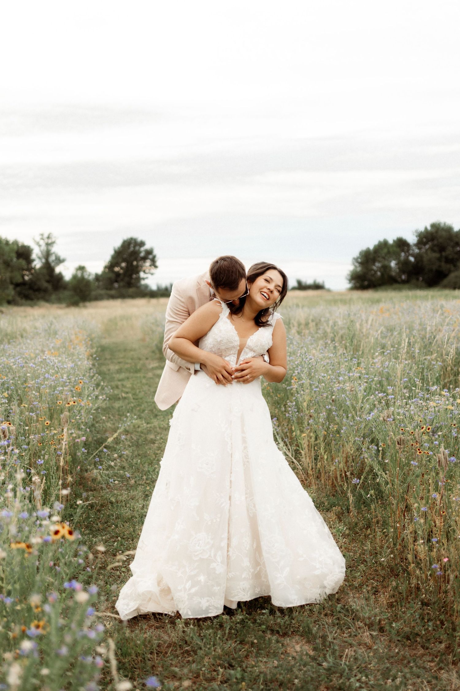 Two people in white dresses share a joyful embrace while standing in a wildflower meadow on a summer day.