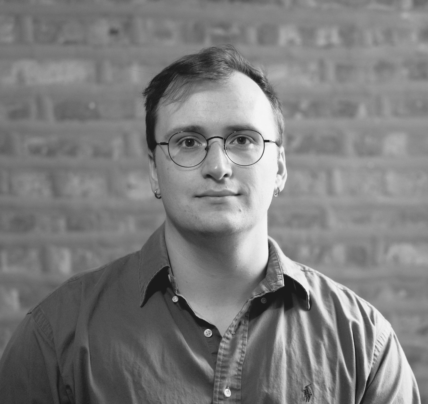Black and white portrait of a young man wearing glasses and a collared shirt, posed against a brick wall background.