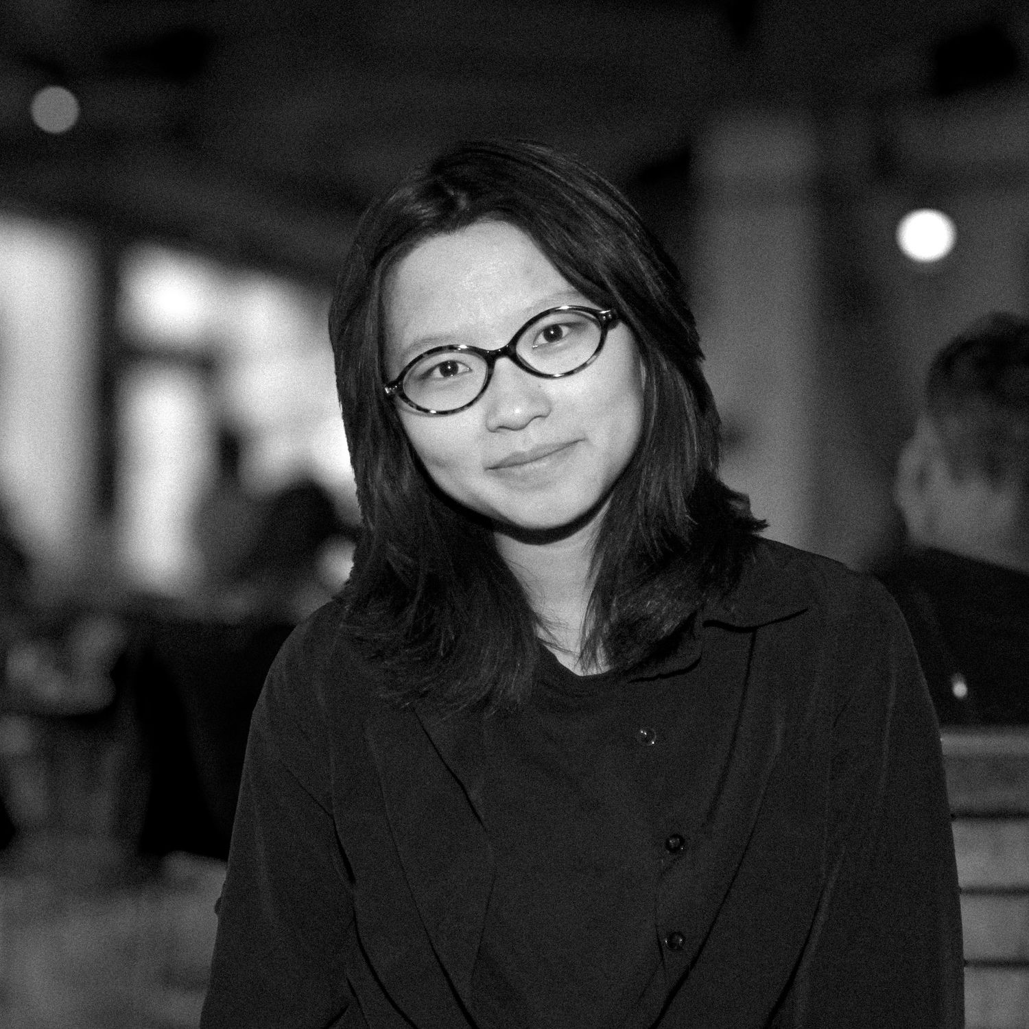Black and white portrait of a young Asian woman wearing glasses and a dark jacket, smiling softly indoors.