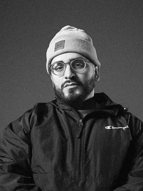 Black and white portrait of someone wearing glasses, a beanie hat and Champion jacket against dark background.