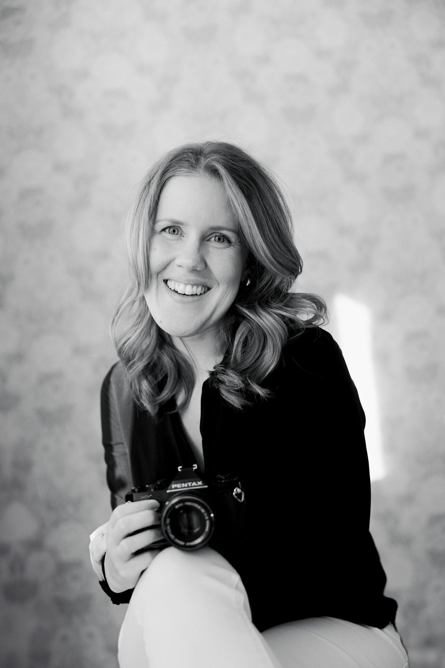 A black and white portrait of a photographer wearing a dark sweater while holding a camera and smiling.