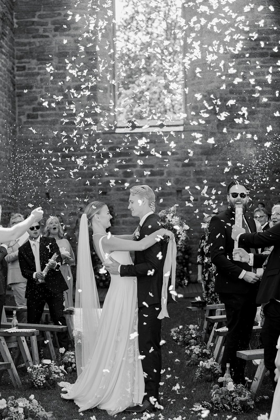A black and white wedding celebration with flower petals raining down on the couple during an outdoor ceremony.