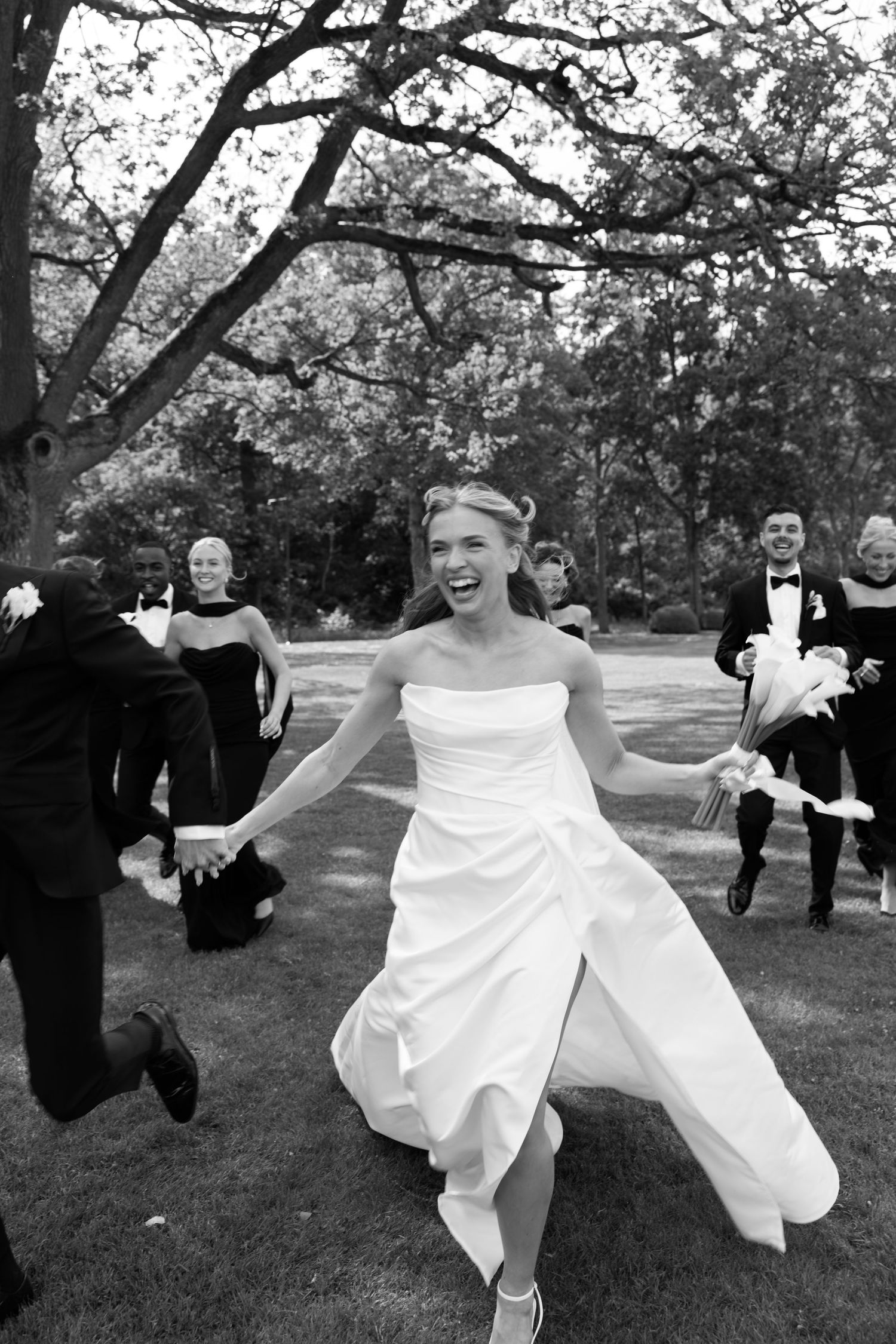 A wedding party jumps and dances playfully on a lawn beneath large tree branches in black and white.