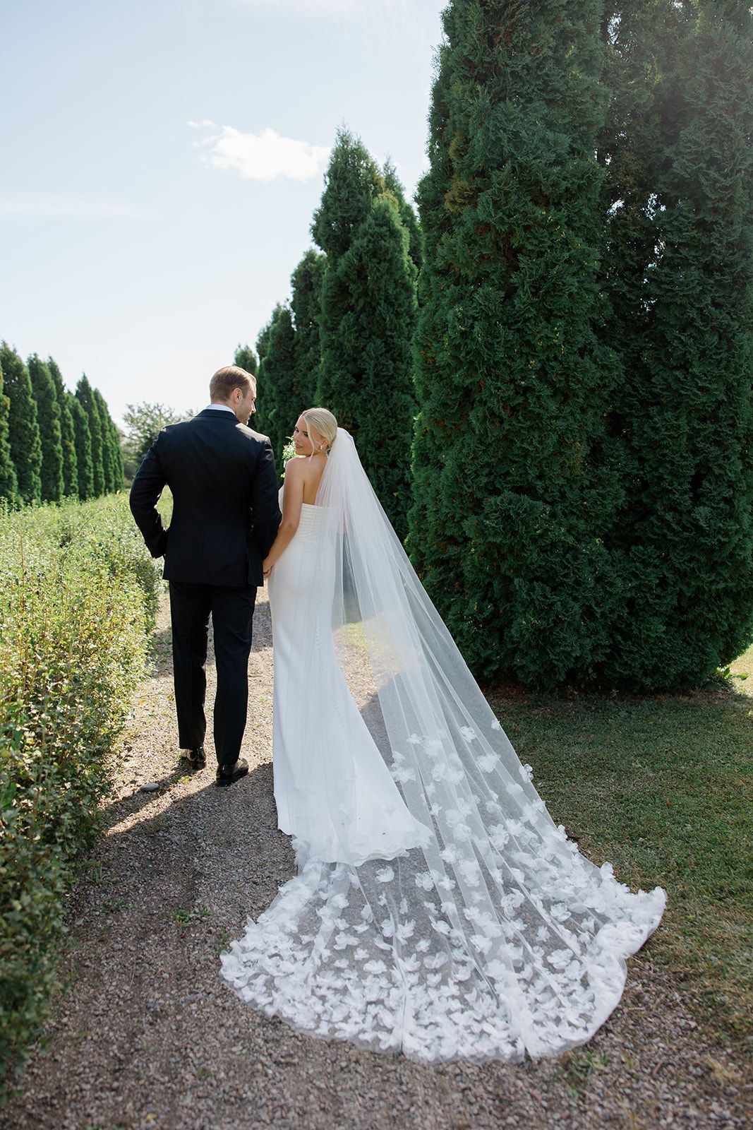 A couple in wedding attire walks along a garden path lined with tall cypress trees on their special day.