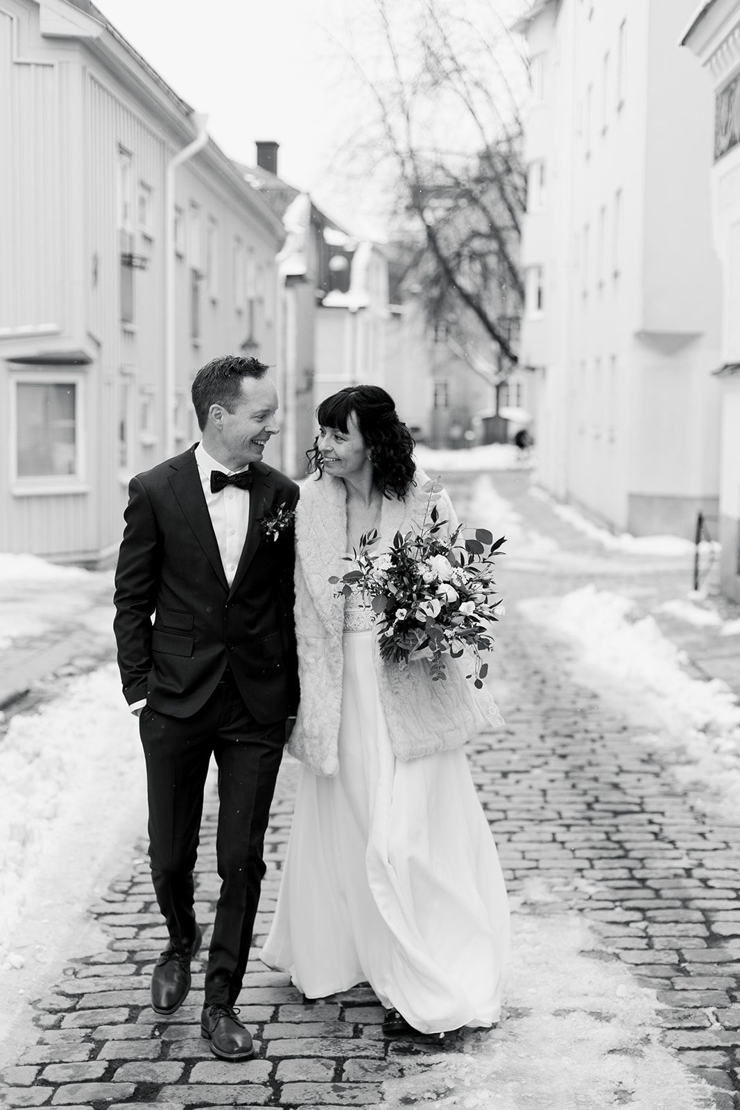 A couple in wedding attire walks together on a cobblestone street in winter with white buildings in the background.