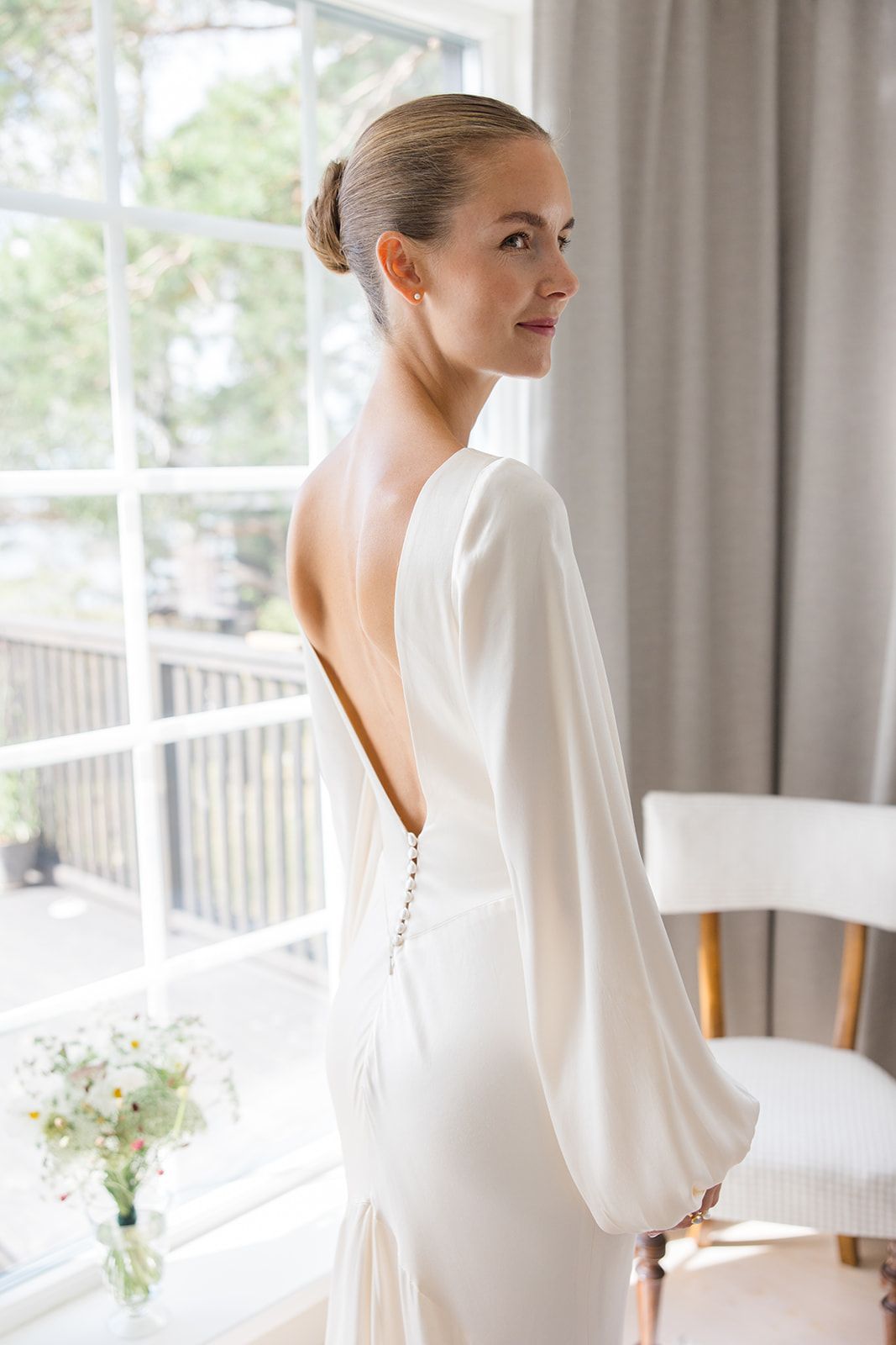 Bride in elegant white backless wedding gown with long sleeves, standing by a bright window, looking over her shoulder.