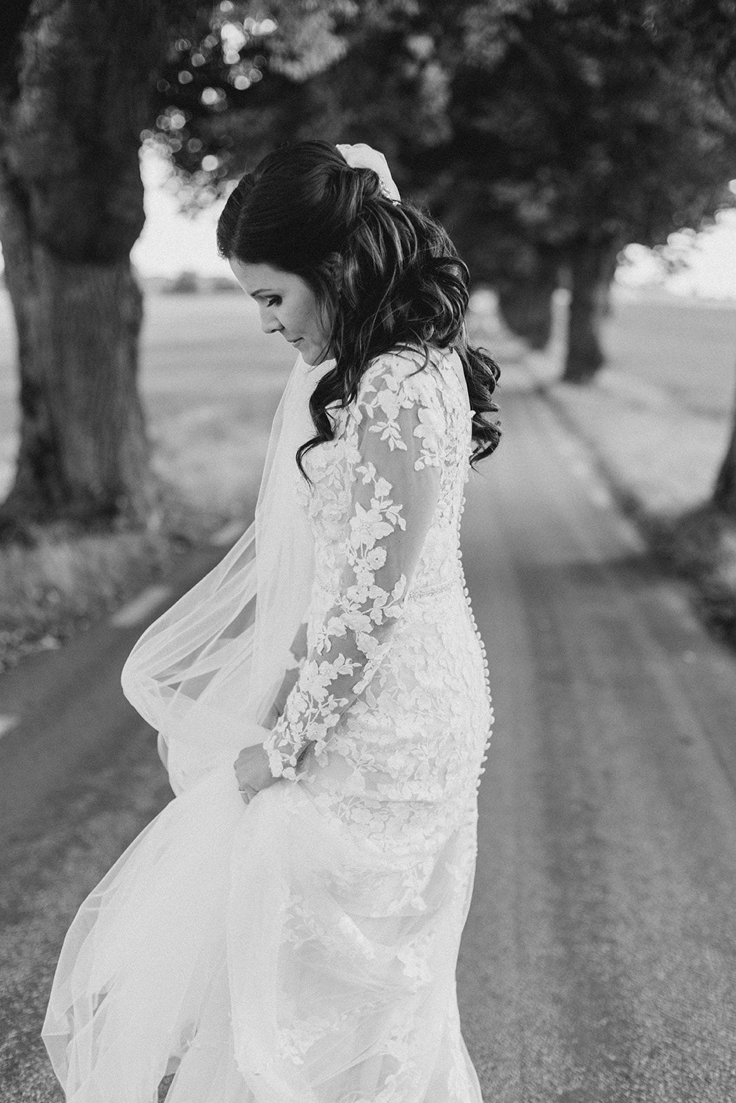 A bride in a flowing lace veil walks along a tree-lined path in black and white photography.