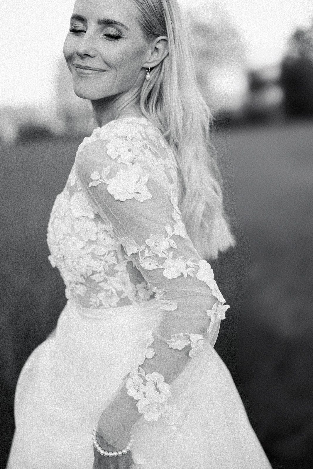 Black and white portrait of a bride in a delicate floral lace wedding gown with sheer sleeves smiling with eyes closed.
