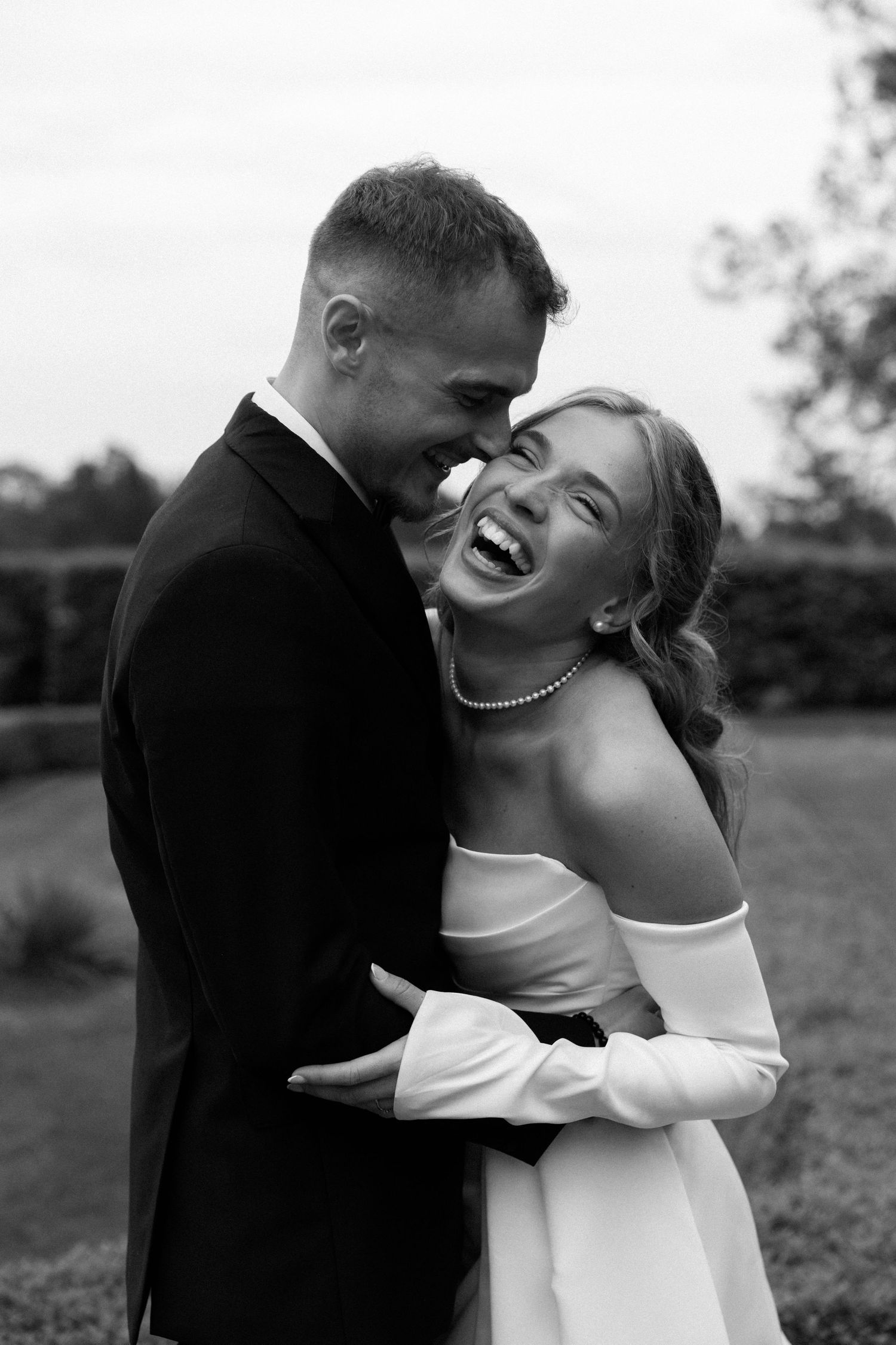 A joyful black and white wedding photo capturing a candid moment of laughter between newlyweds outdoors.