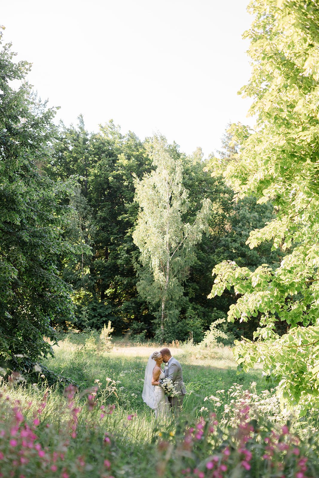 A couple stands together in a sunlit meadow with purple wildflowers blooming in the foreground and trees in the background.