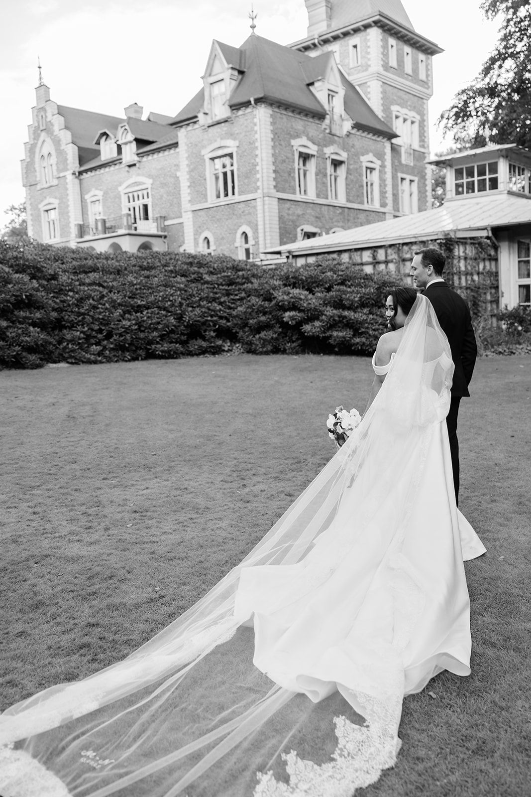 A couple in wedding attire embraces on a grand lawn with a historic stone mansion in the background.