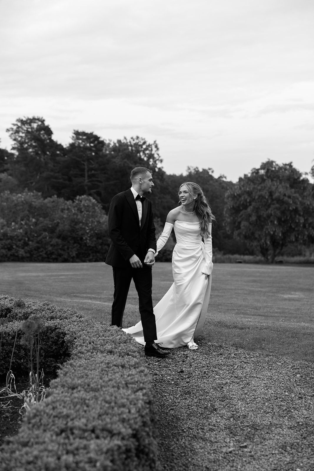 A couple in wedding attire walks together across a grassy hillside in black and white.