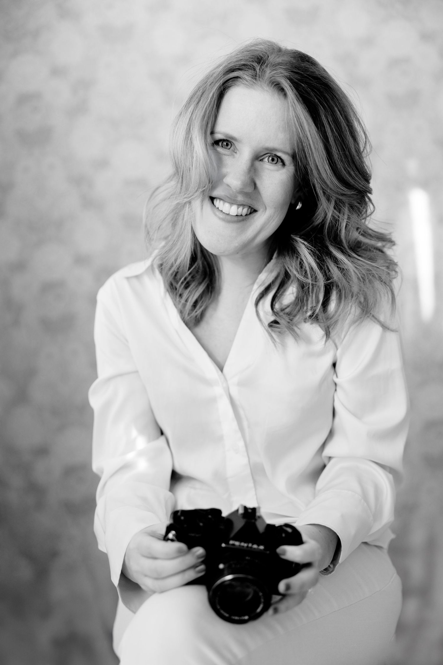 Black and white portrait of photographer holding a camera while wearing a white button-up shirt and smiling warmly.