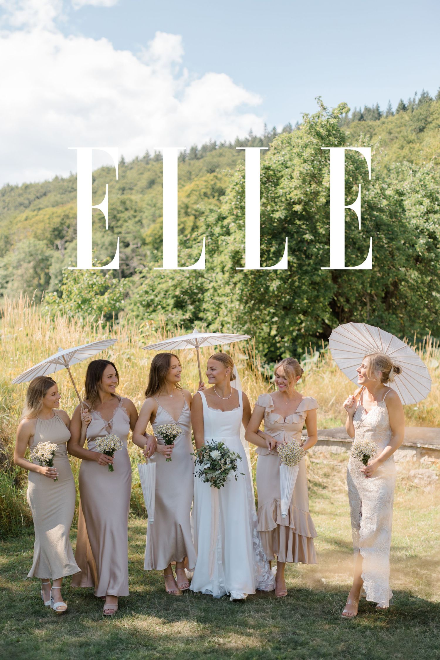 Wedding party in neutral dresses posing outdoors with parasols against a scenic hillside backdrop.