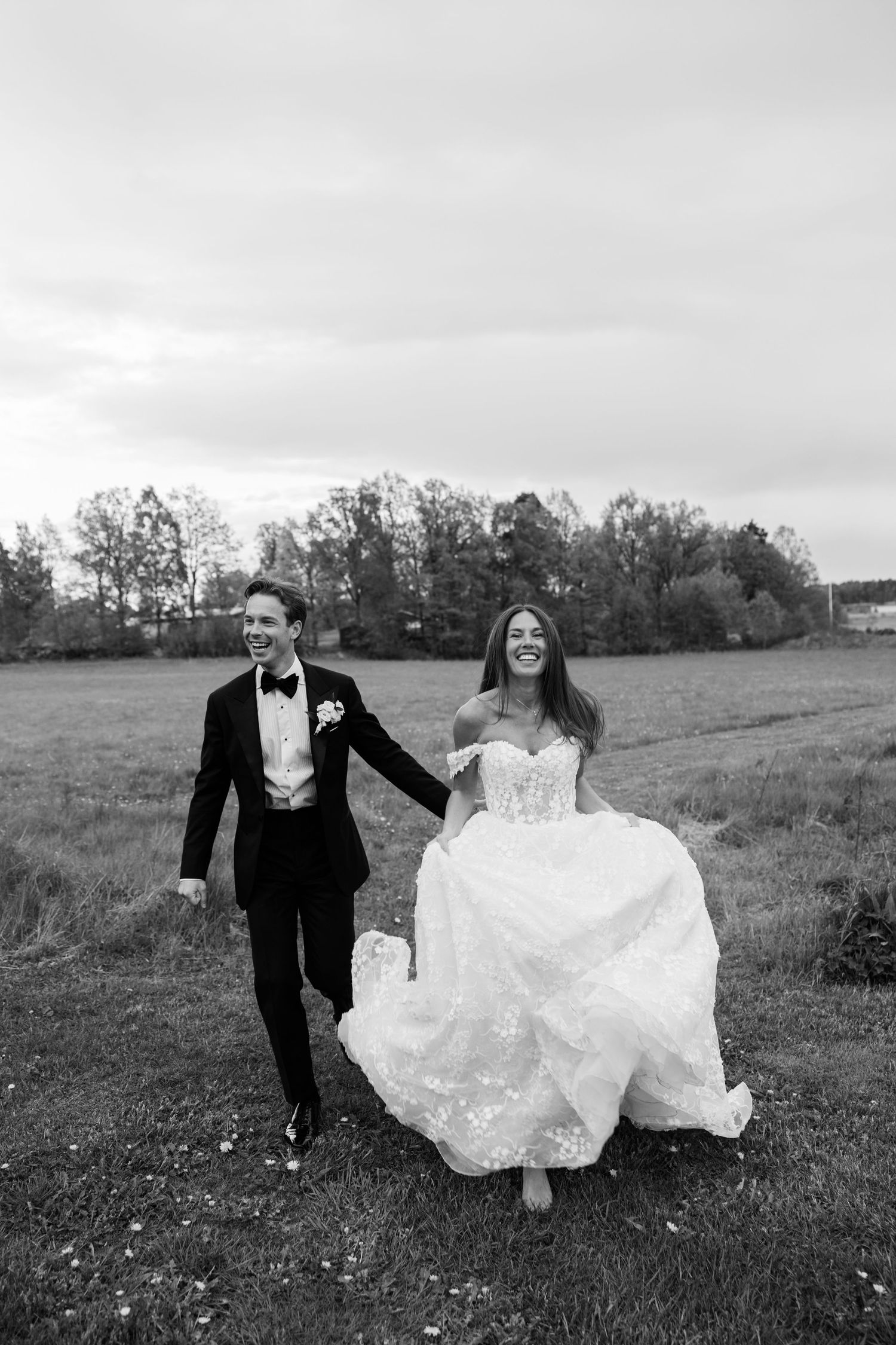Black and white photo of smiling bride and groom holding hands running through a field on their wedding day.