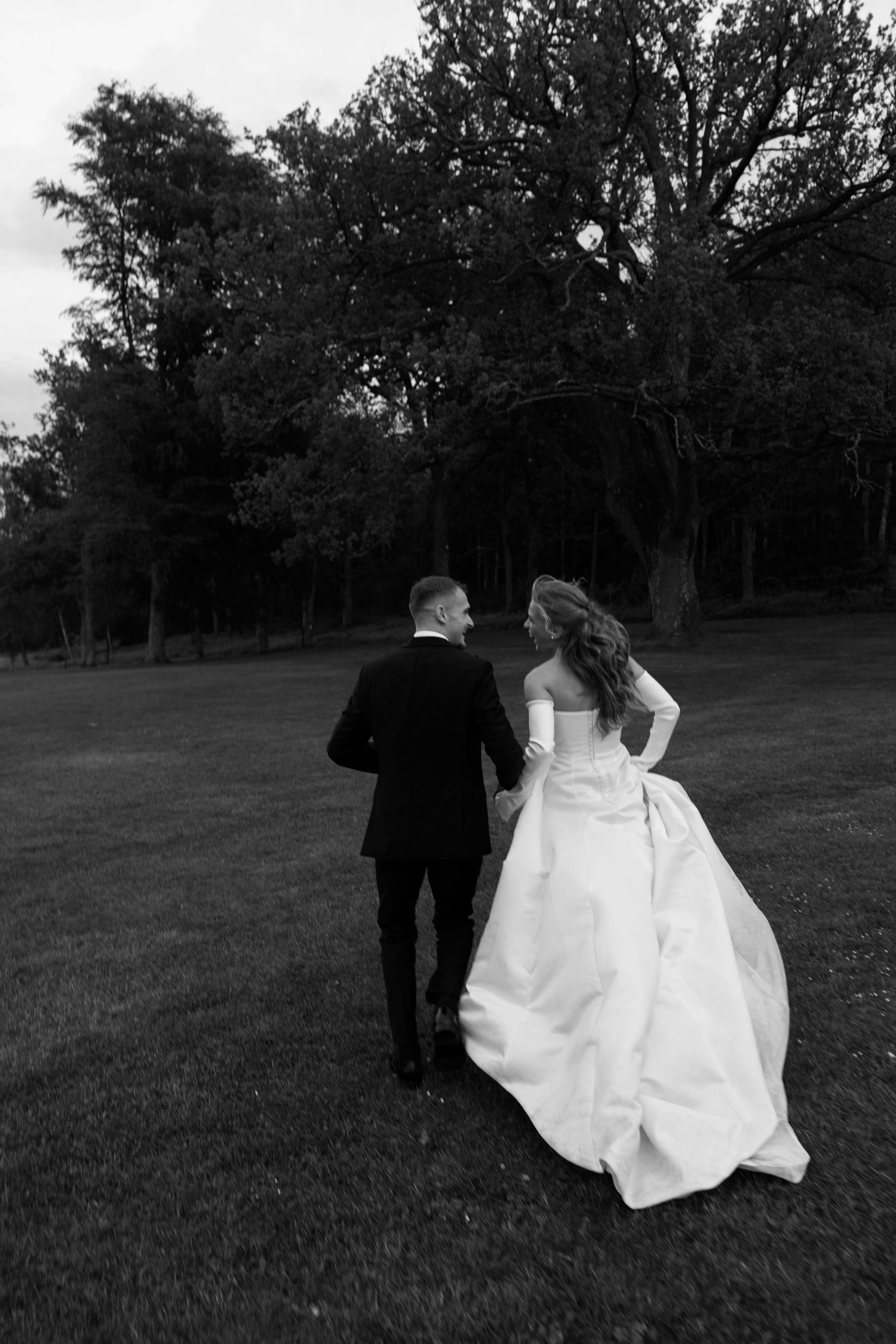 Black and white photo of newlyweds walking hand in hand across a grassy lawn with trees in the background.