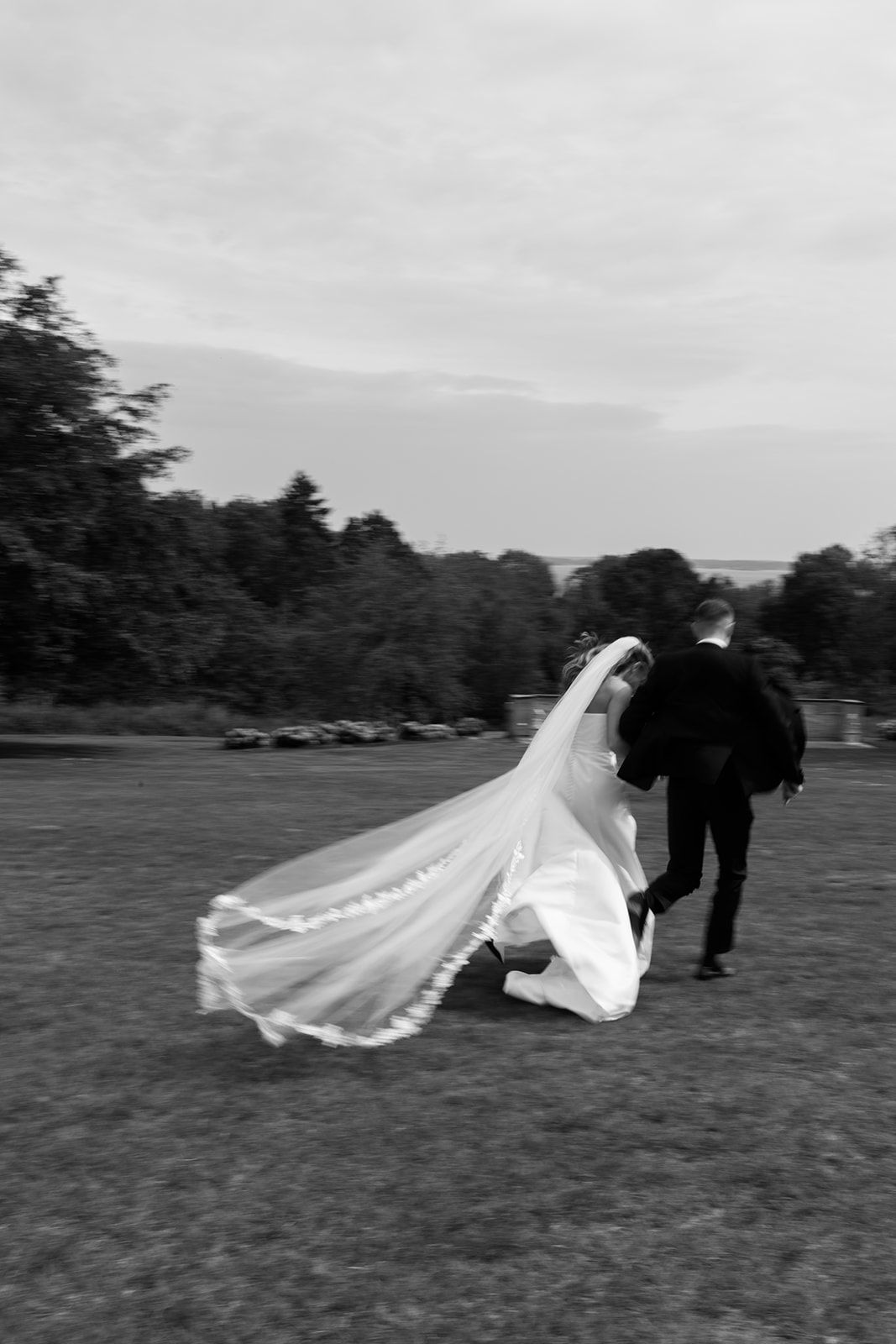A bride's veil flows dramatically in the wind as a couple poses on a grassy field in black and white.