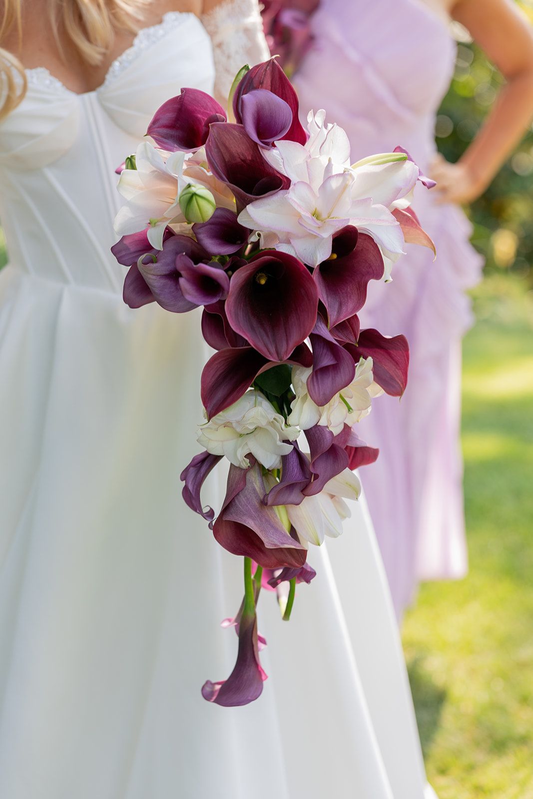 Bride holding cascading wedding bouquet of deep purple calla lilies and white flowers outdoors.