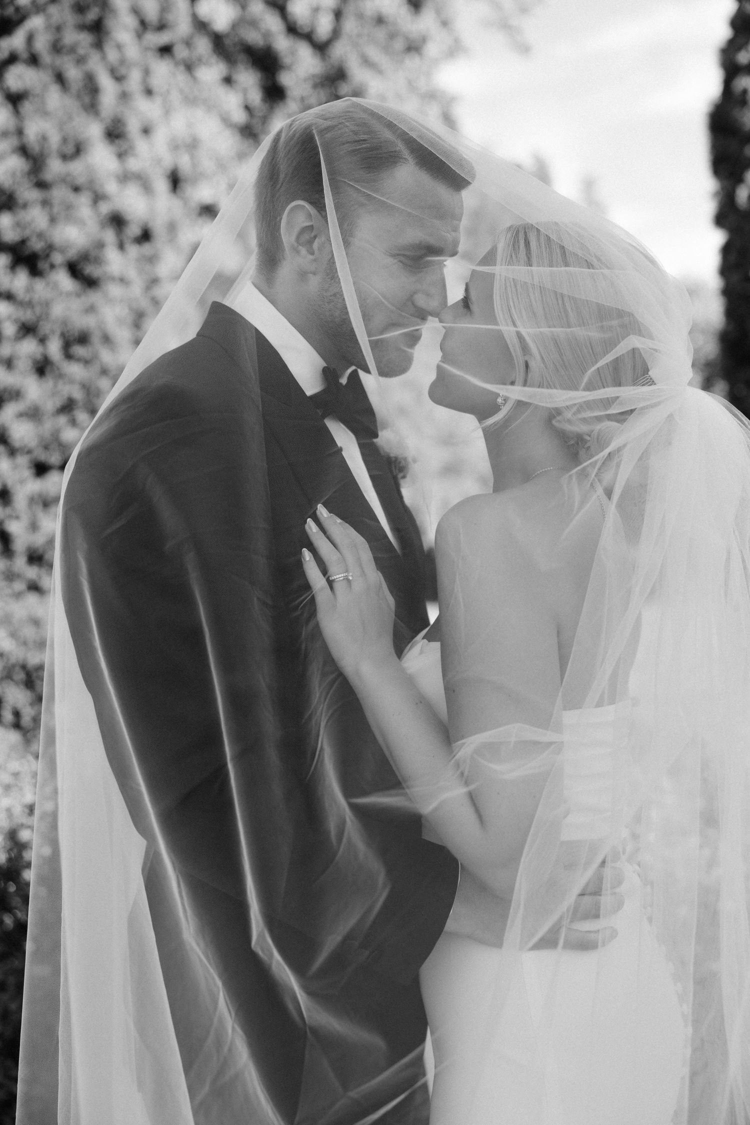 A black and white portrait captures an intimate moment between newlyweds beneath a flowing veil.