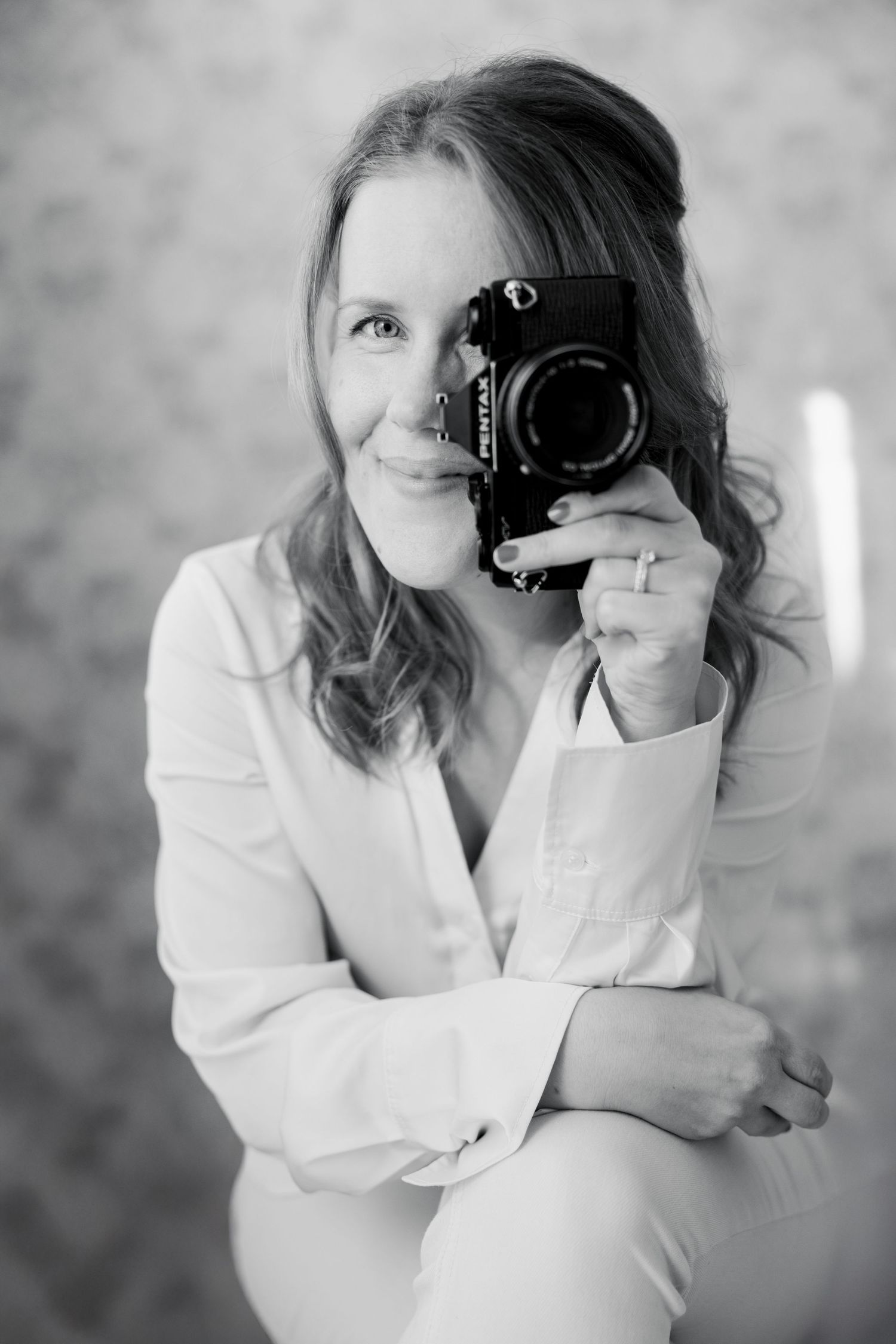 Black and white portrait of a photographer in white attire holding a professional camera and smiling at the lens.