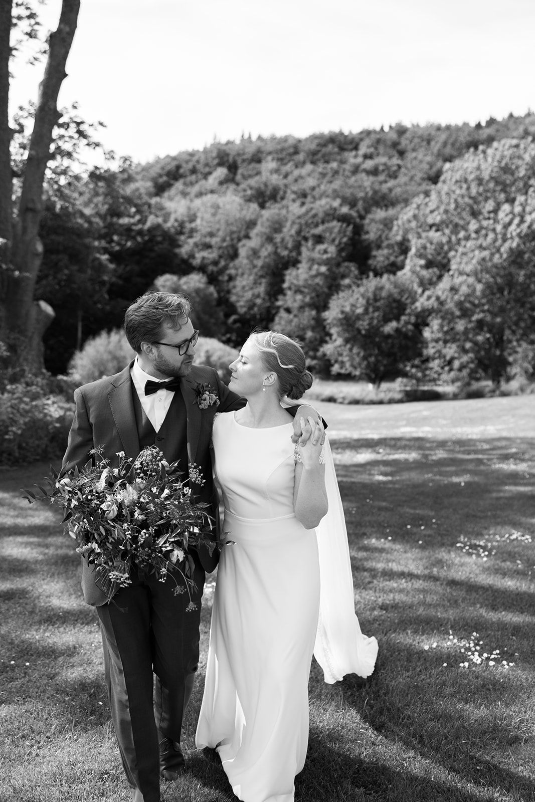 Black and white wedding photograph showing couple walking together through a sunny meadow with trees in background.
