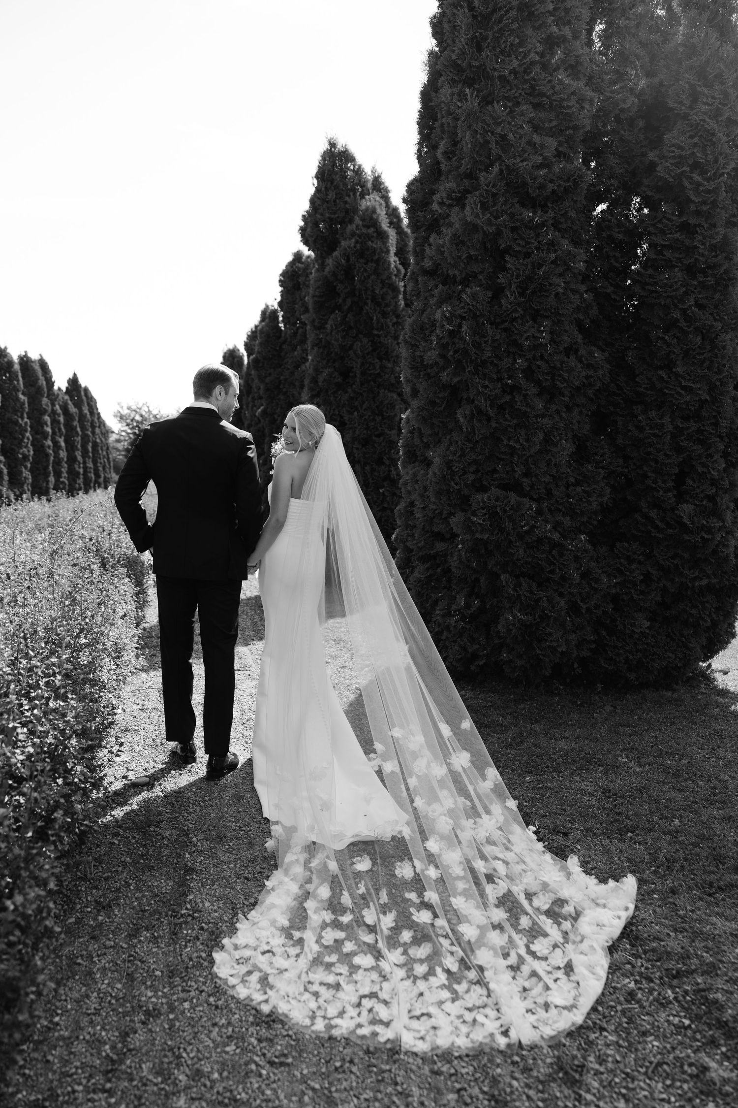 A black and white photo of a couple in wedding attire walking along a path lined with tall cypress trees.
