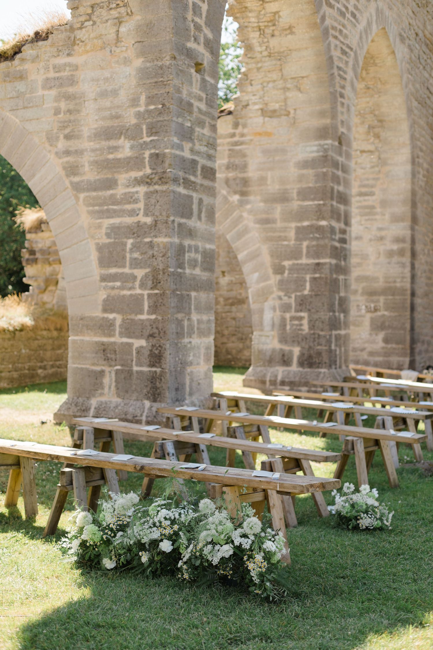 Rustic wooden benches arranged for outdoor seating against stone church walls with white floral arrangements on grass.