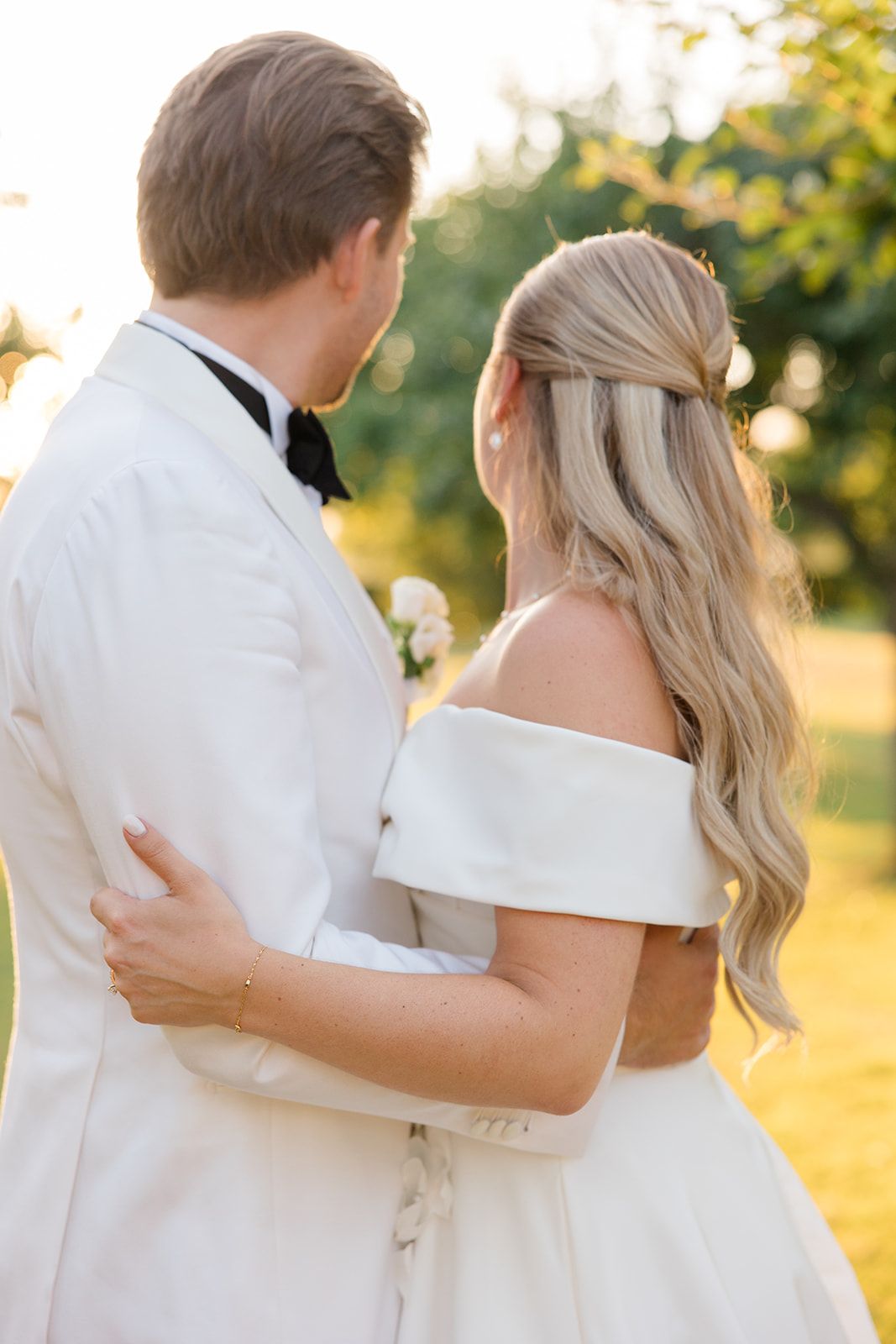 A couple in white attire embraces while walking outdoors during golden hour sunlight.