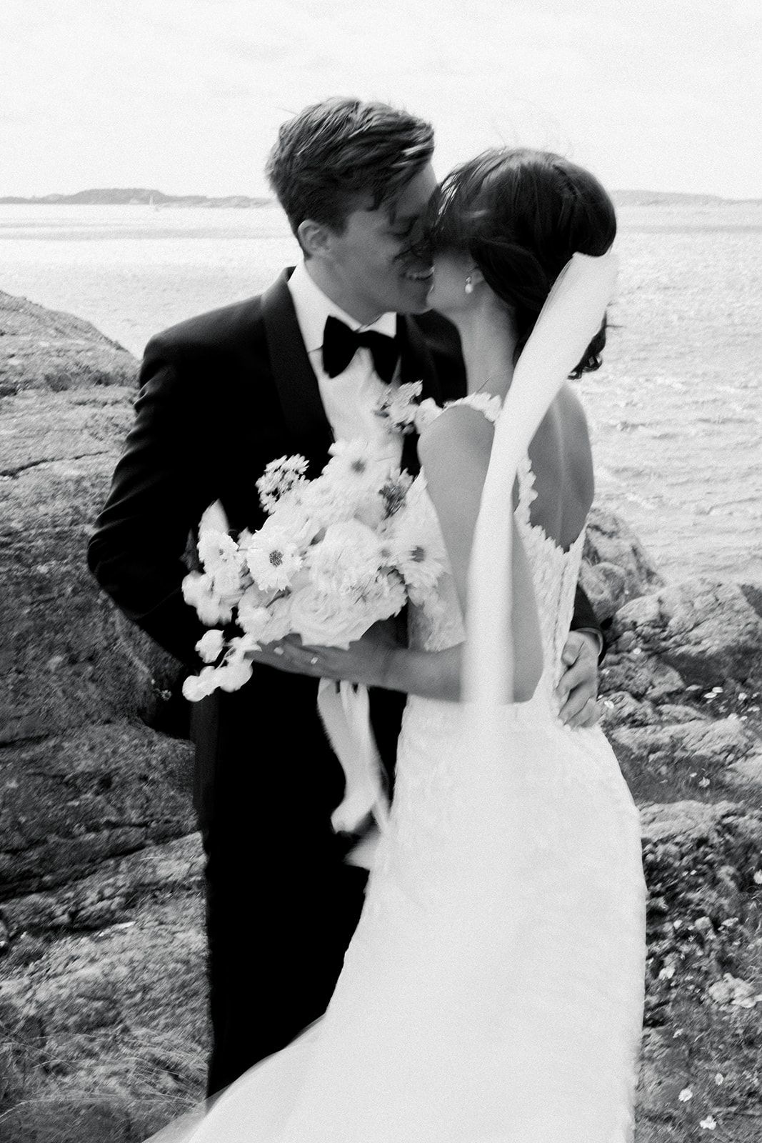 A romantic black and white wedding photo of a couple kissing on rocky coastal cliffs with a bouquet of white flowers.
