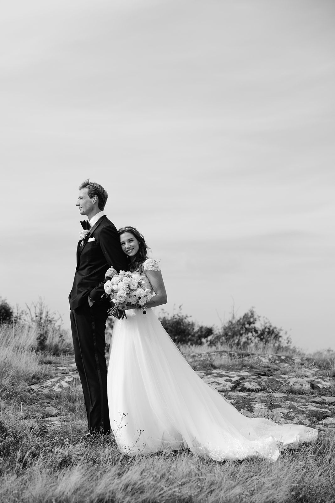 Black and white wedding photo of a couple standing on a hilltop, with the bride holding a bouquet of flowers.