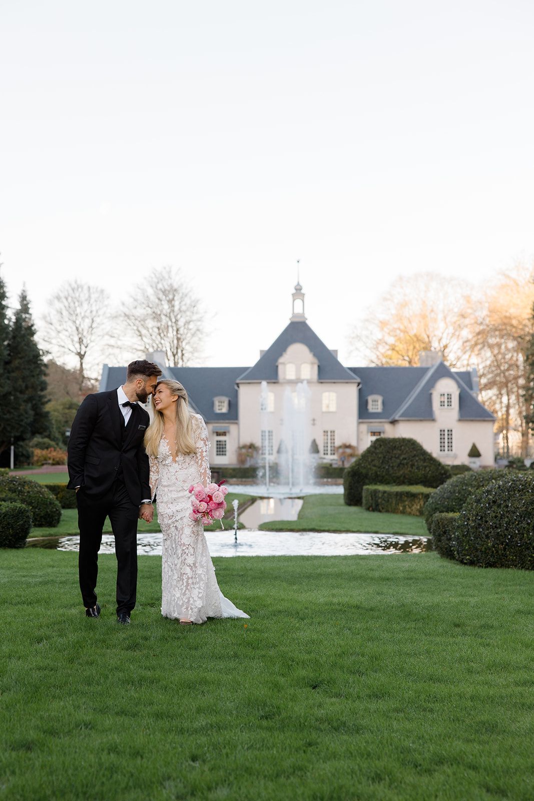 A couple shares a romantic moment on manicured grounds with an elegant white mansion and fountain in the background.