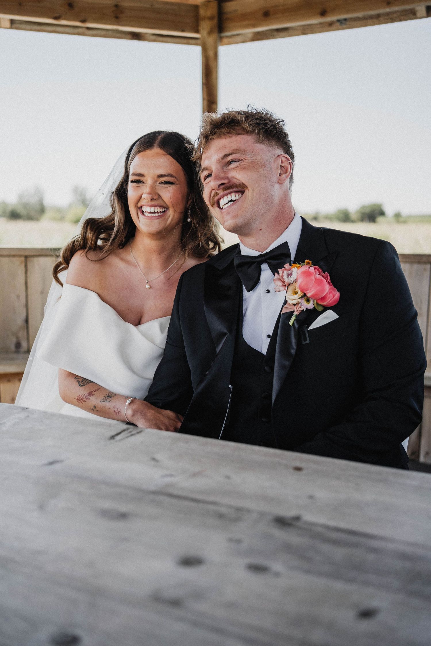 A joyful wedding couple shares a candid laughing moment together while seated at a rustic wooden table outdoors.