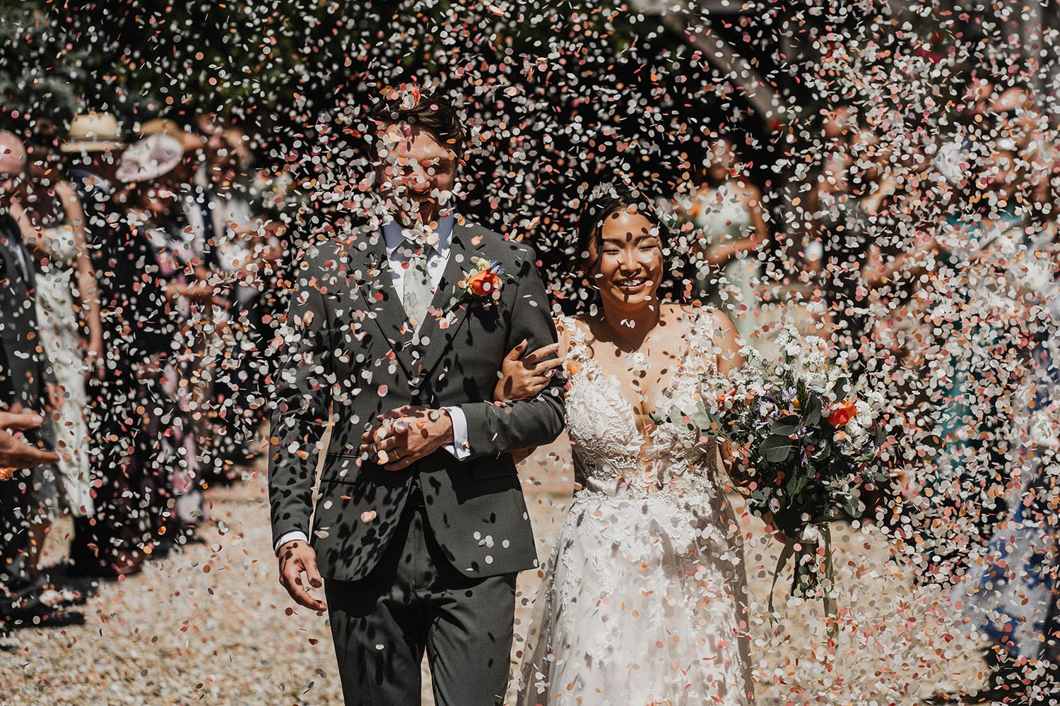 Smiling couple surrounded by a shower of white confetti petals during their outdoor celebration on a sunny day.