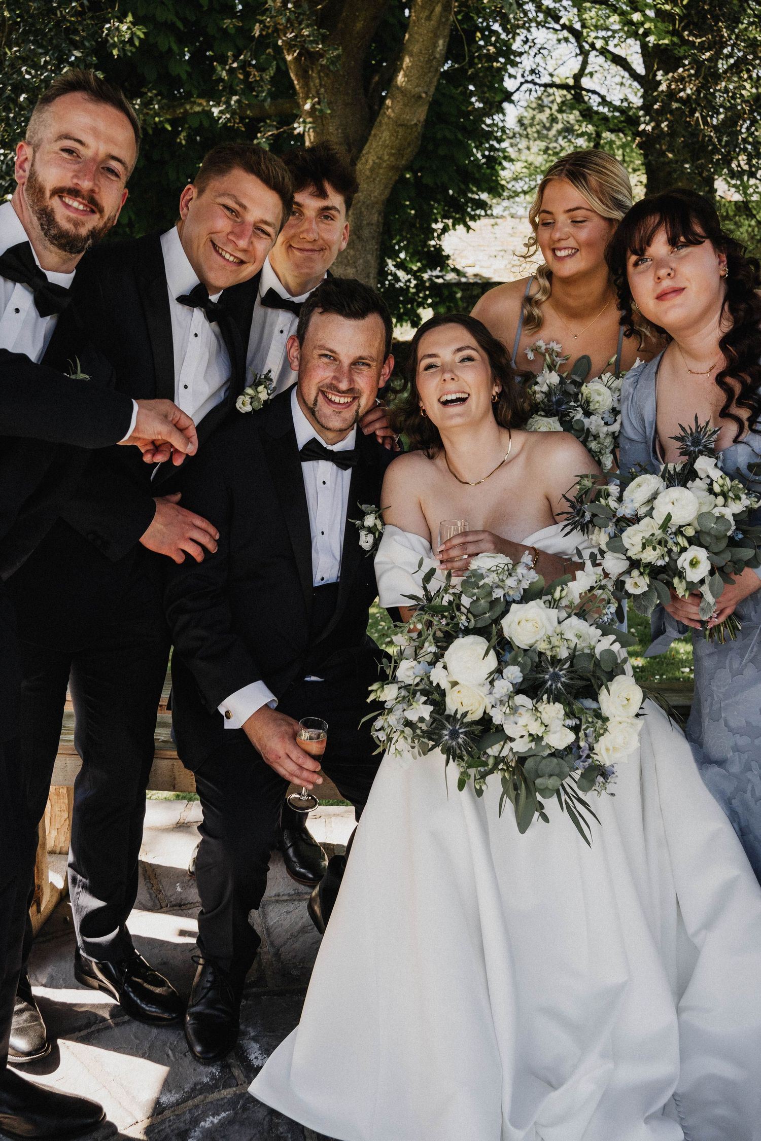 A wedding party poses together outdoors with white floral bouquets, the men in black tuxedos and bride in a flowing white gown.