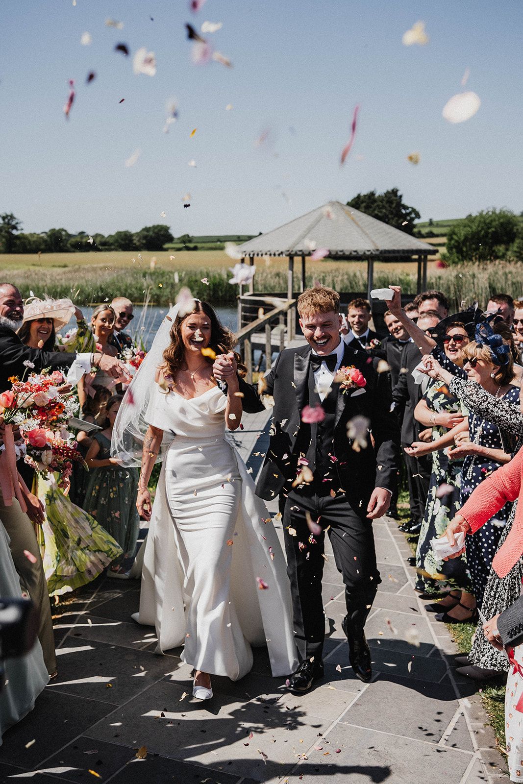 A joyful wedding celebration with confetti being thrown over a couple walking down an outdoor path under a gazebo.