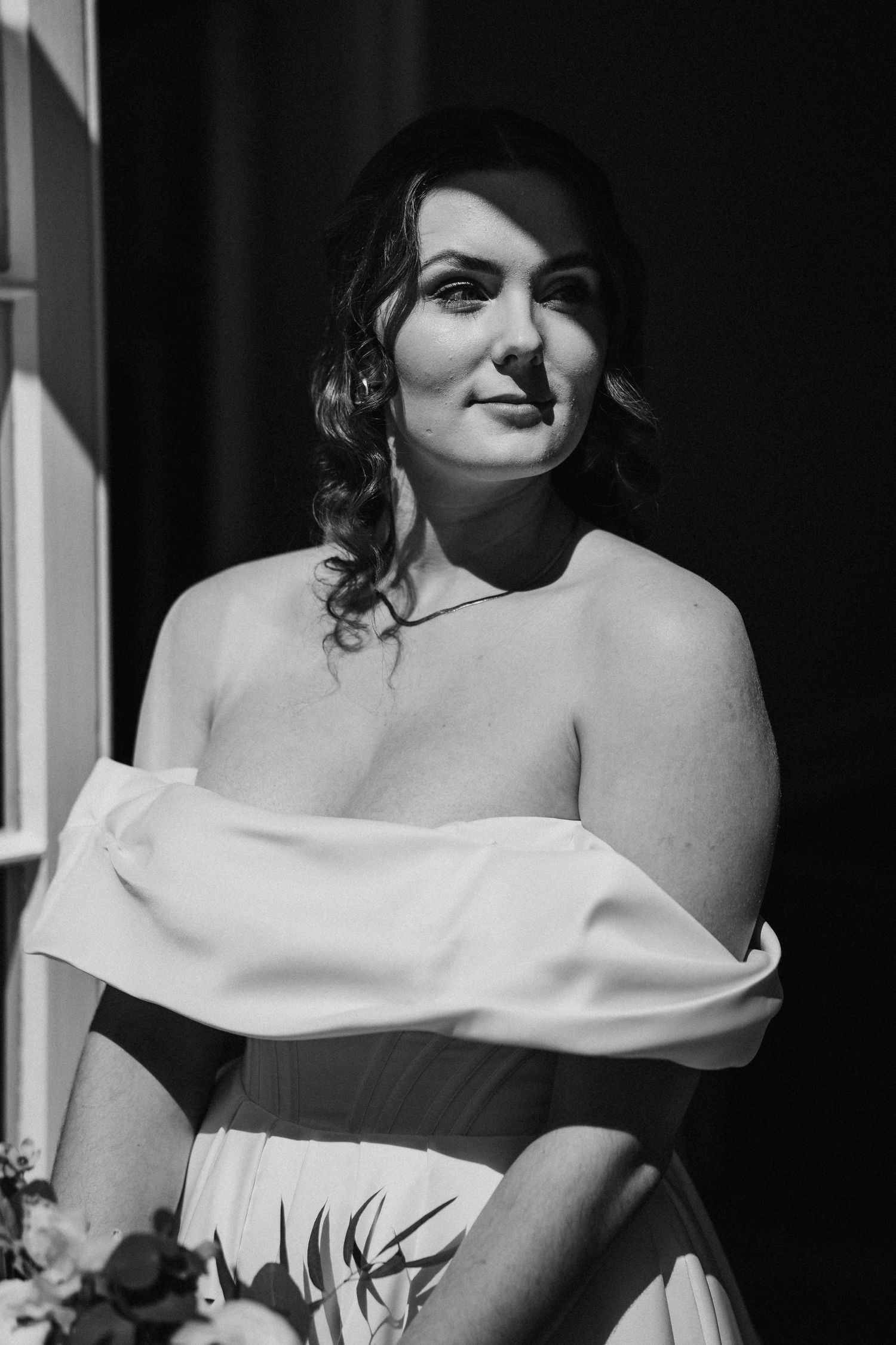 Black and white portrait of someone in an off-shoulder white dress with wavy hair looking thoughtfully upward.