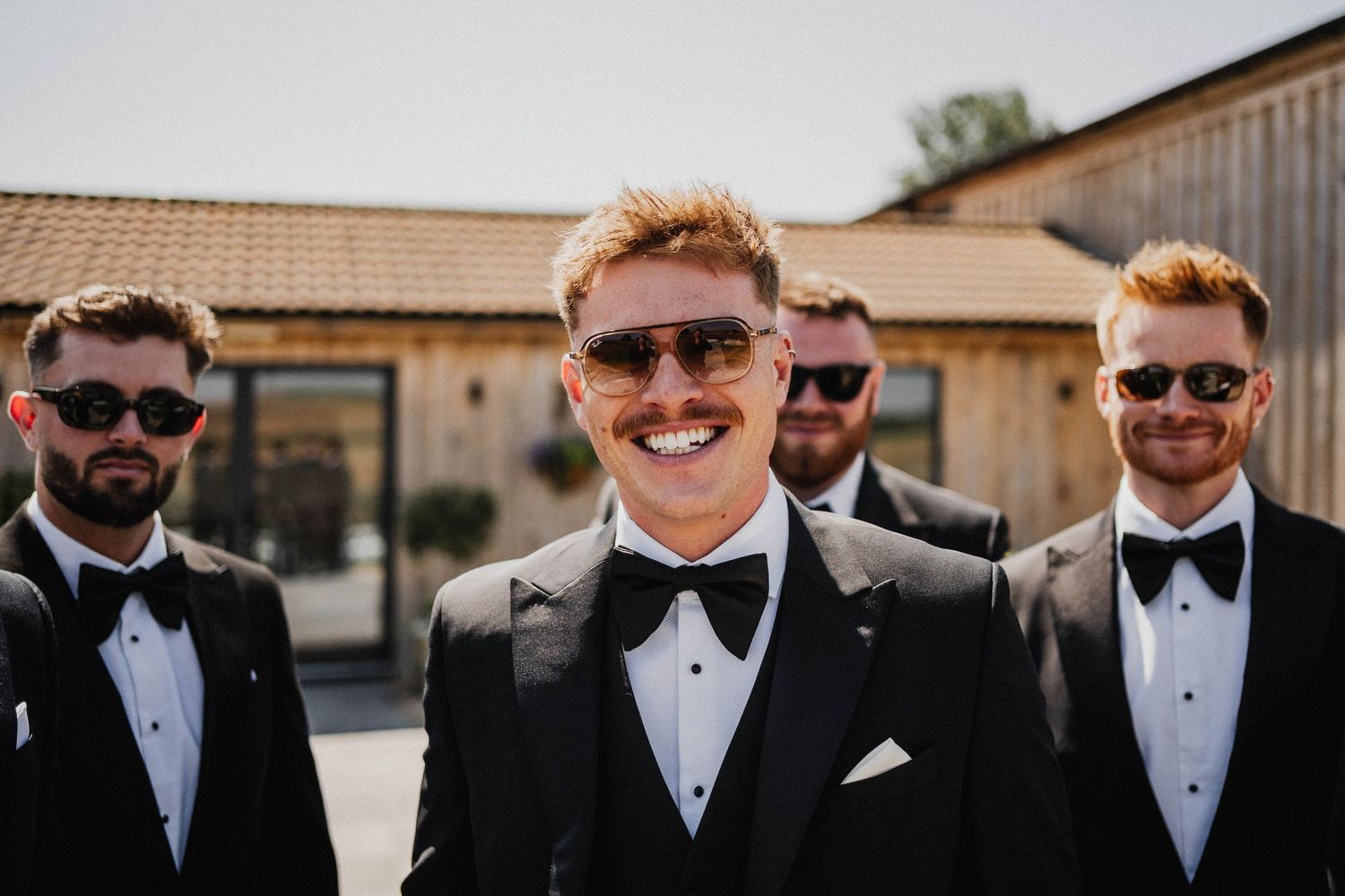 Groomsmen in black tuxedos and sunglasses pose together outdoors at a formal wedding event.