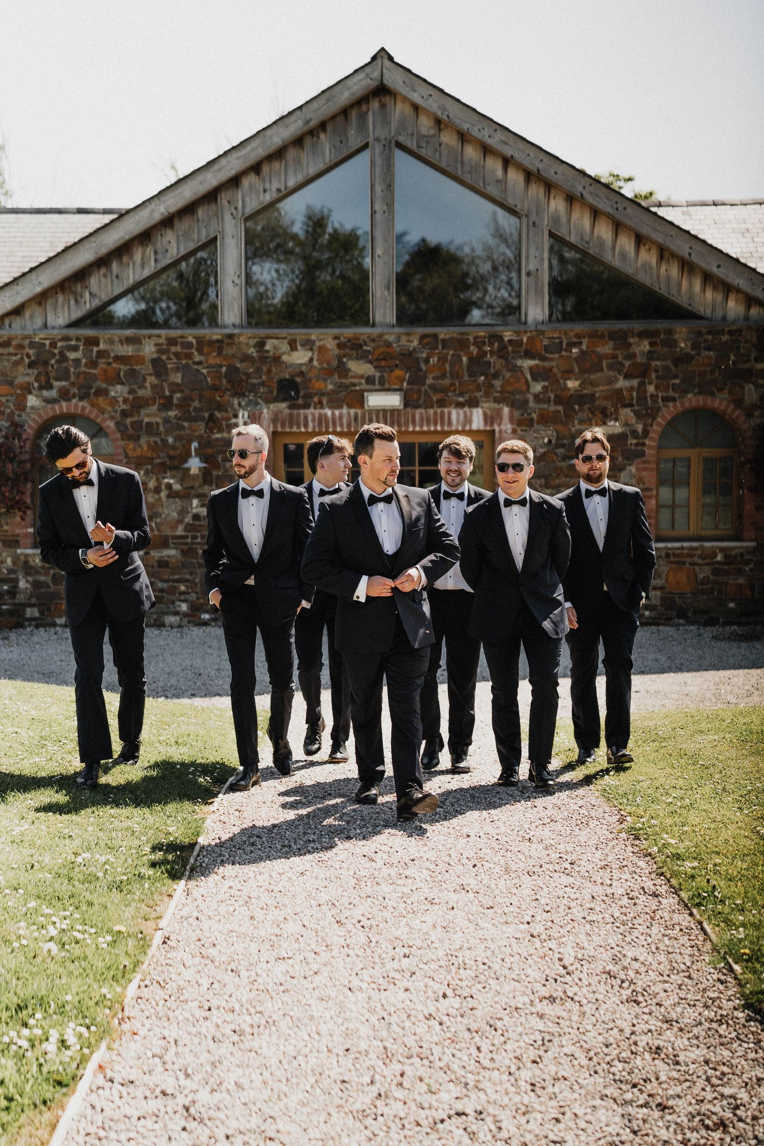 A group wearing black tuxedos and bowties walks along a gravel path outside a rustic stone building with glass windows.