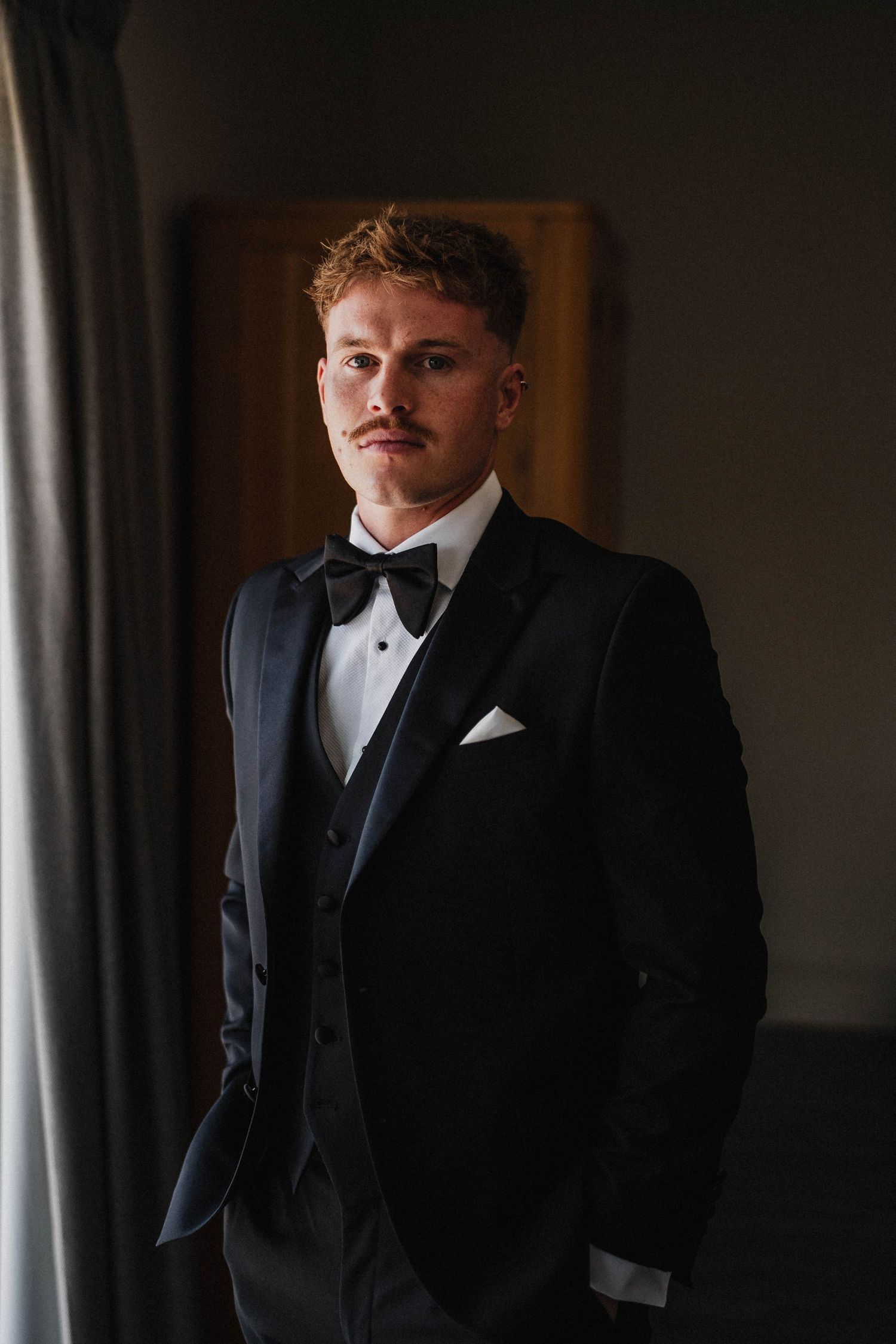 Portrait shots of a person in a formal black tuxedo and bowtie standing near window light in moody lighting.