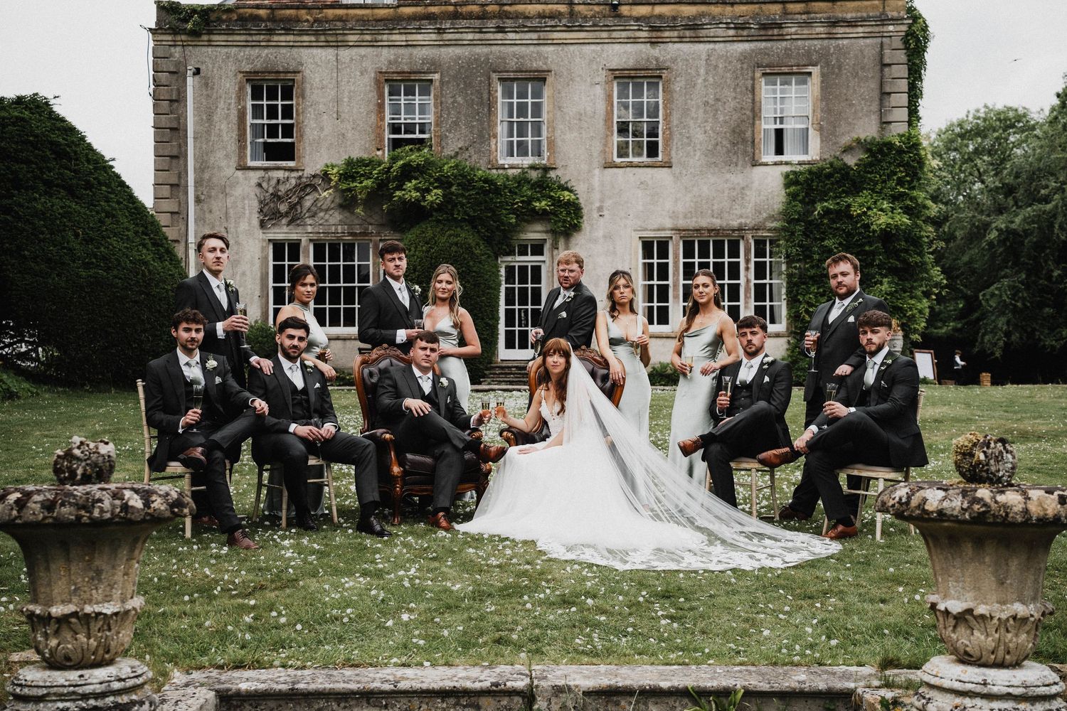 Wedding party sits on stone wall outside historic mansion during summer celebration.
