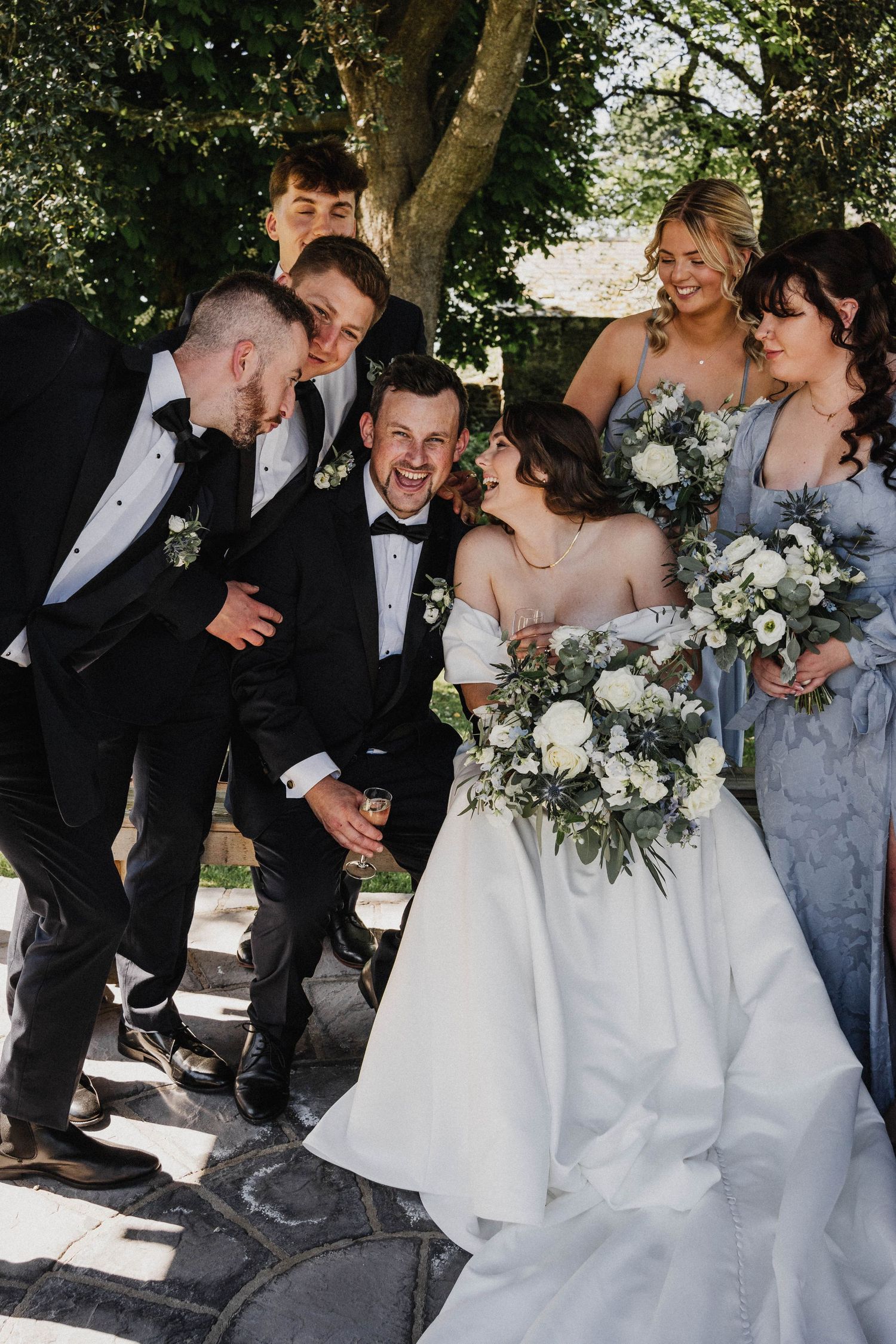 A joyful wedding party shares a candid moment outdoors in formal attire, with bridesmaids in blue dresses and white bouquets.