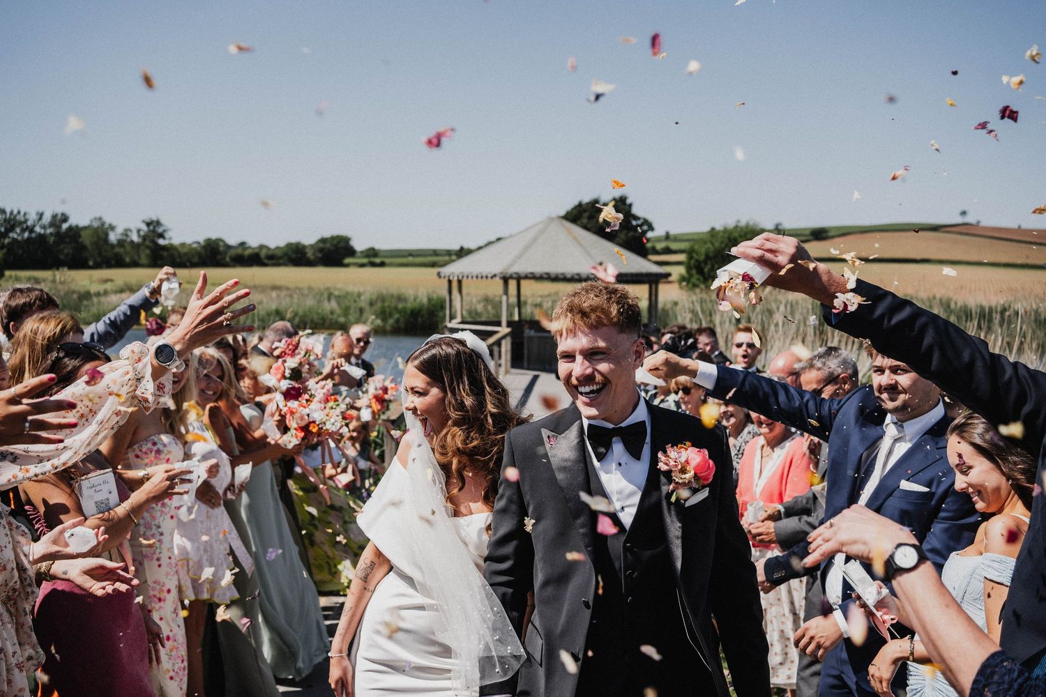 Wedding guests shower confetti on a newlywed couple during an outdoor celebration.