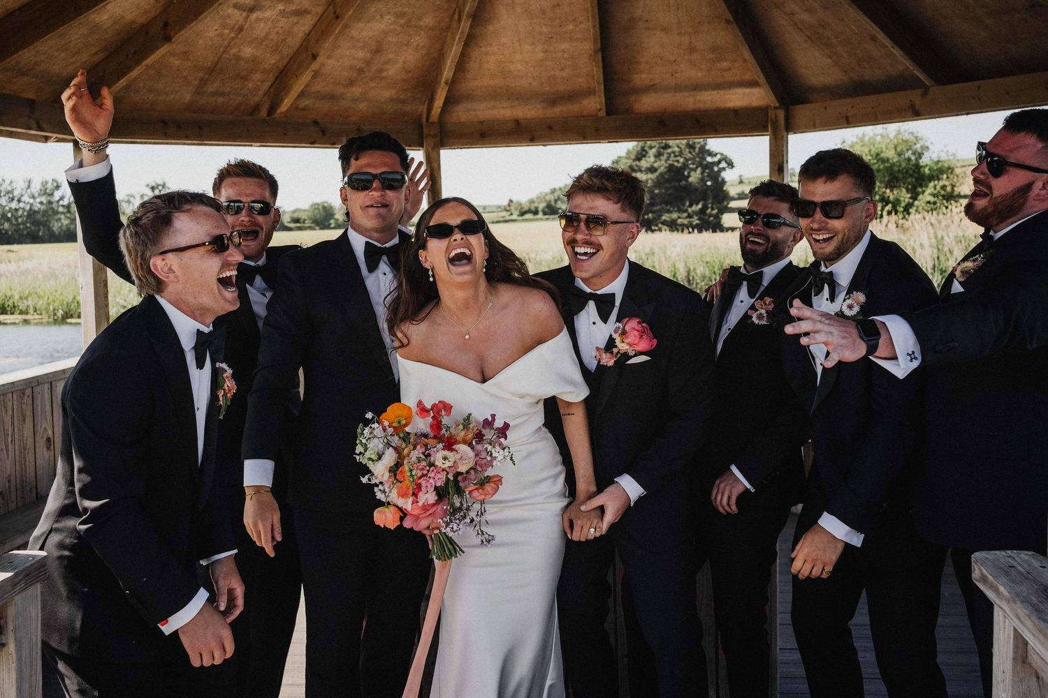 A wedding party gathers outdoors under a rustic gazebo, sharing laughter and smiles on the special day.