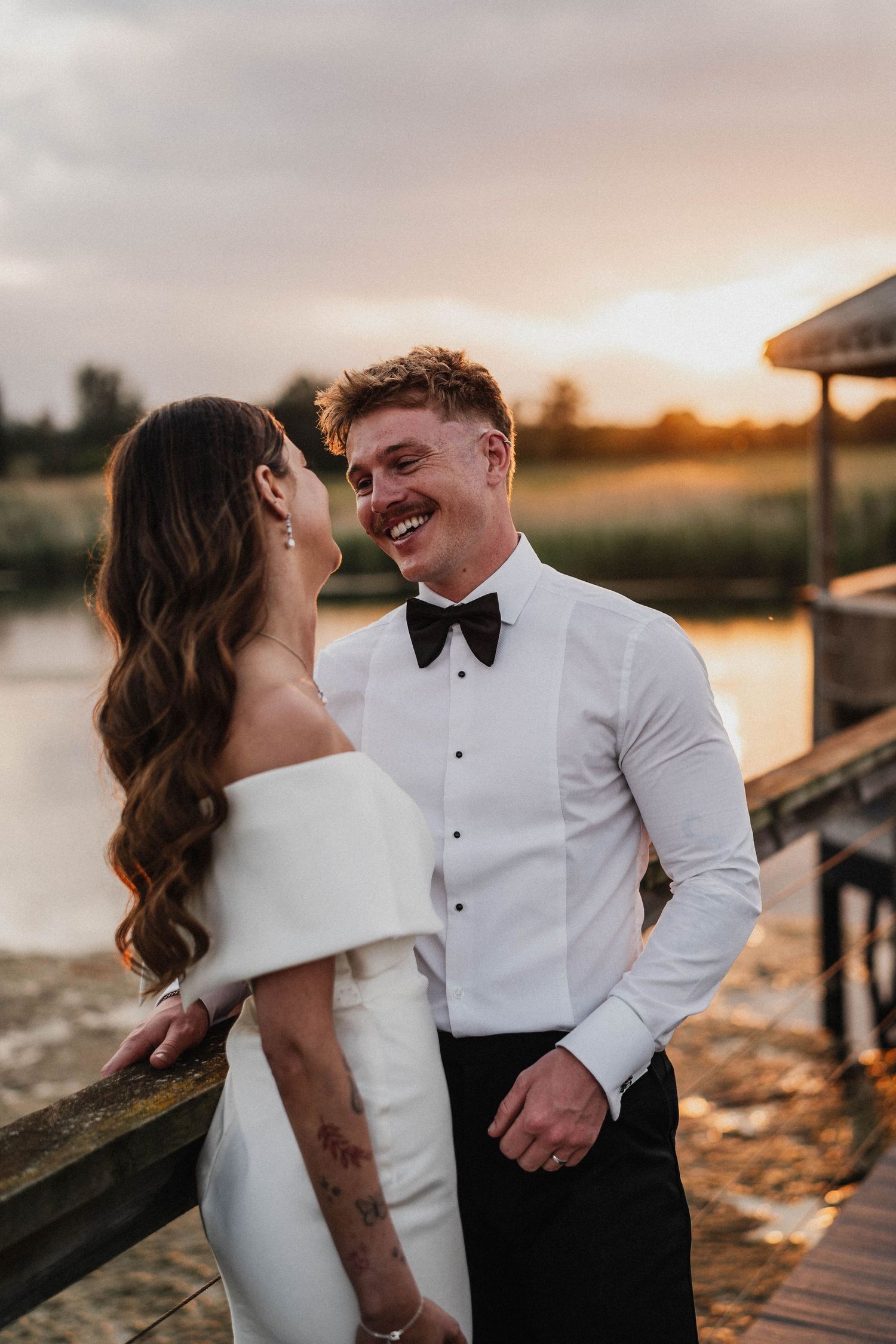 A romantic couple in formalwear share a tender moment at sunset on a wooden dock.